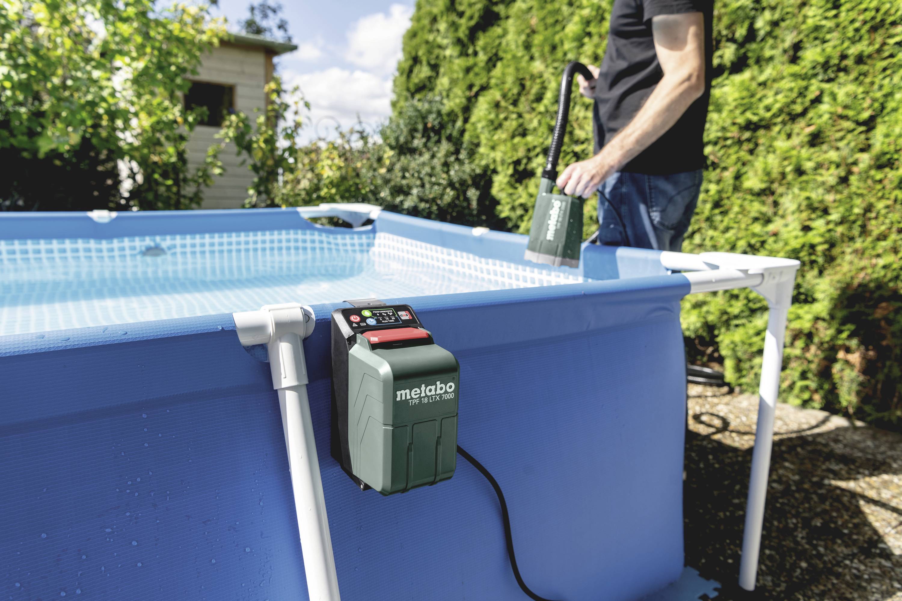 A large, blue, rectangular outdoor pool. A person is holding a pump over the pool. A Metabo pump is mounted in the foreground.