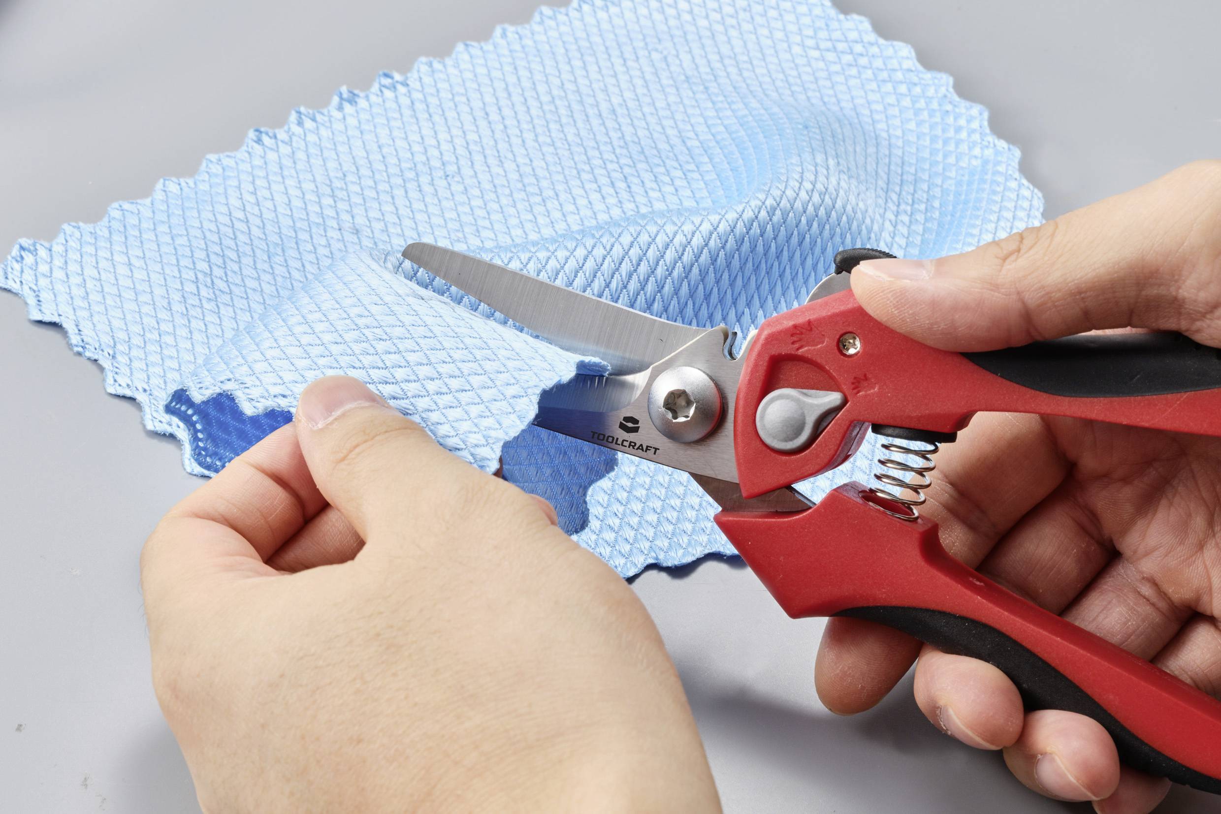 'A person is cutting blue fabric with red scissors on a grey background.'