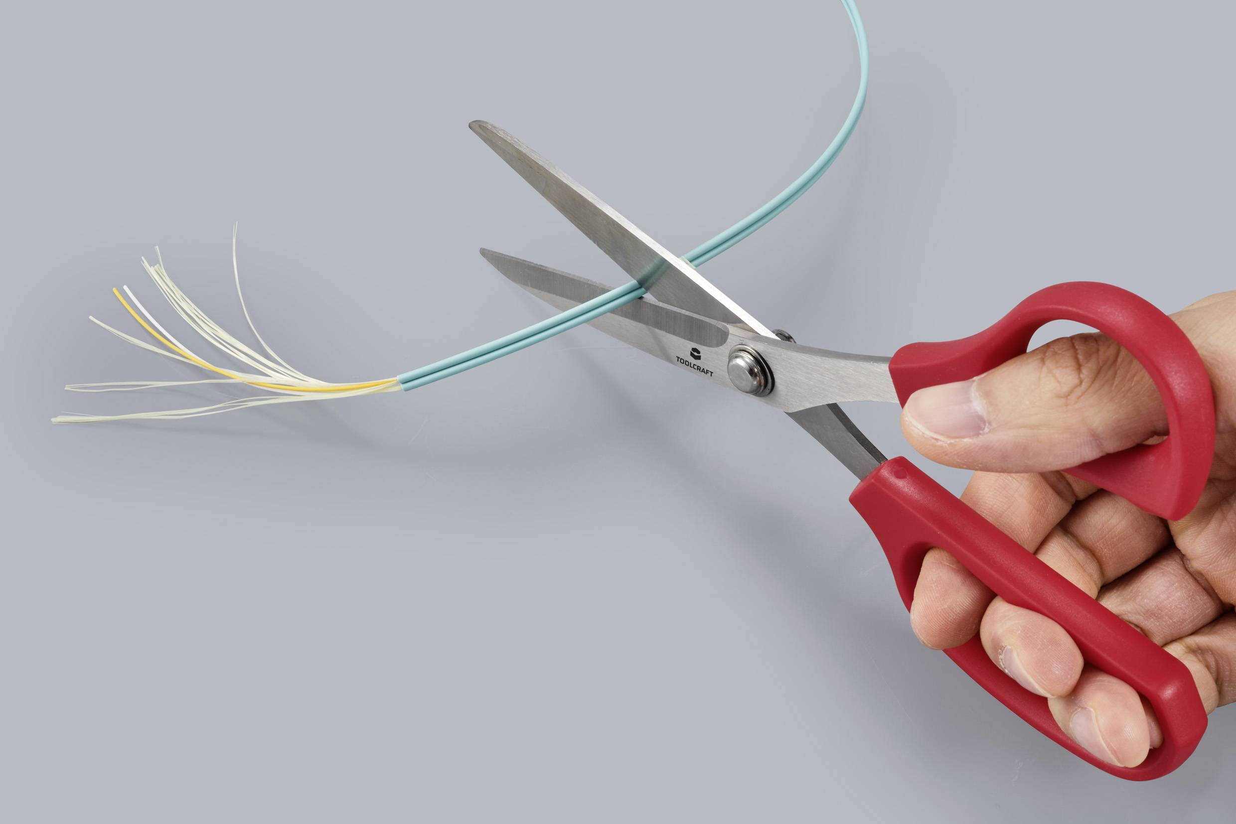 A red pair of scissors cuts a thin blue cable with yellow and white fibres, held in a hand, against a grey background.