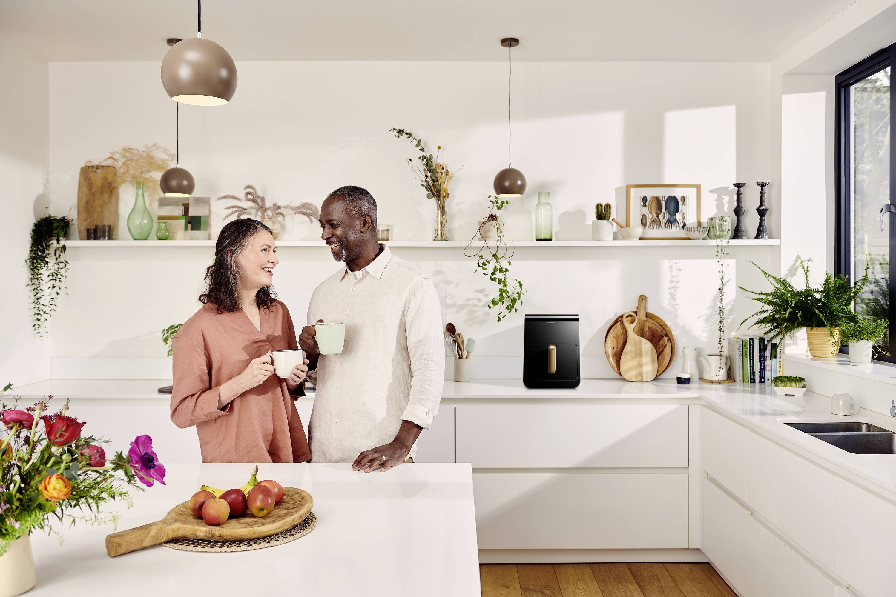 A man and a woman are standing smiling in a modern kitchen, both holding mugs, surrounded by plants and decorations.