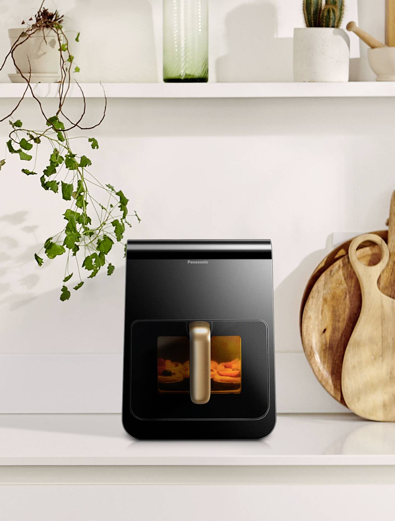 Compact modern mini oven on a kitchen worktop, surrounded by wooden boards and plants, with a glossy black surface.