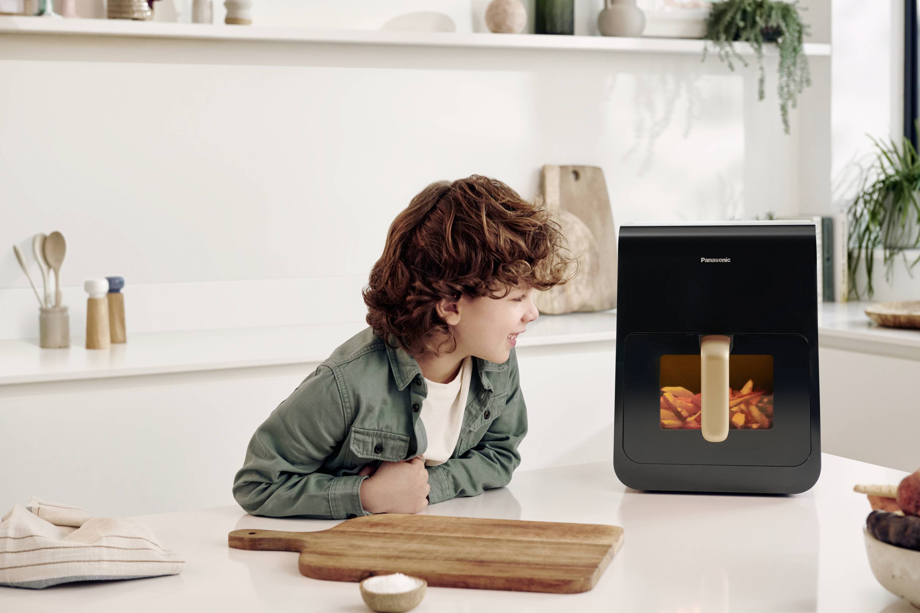 A child gazes in fascination at an air fryer preparing chips. The kitchen is bright and modern in its design.