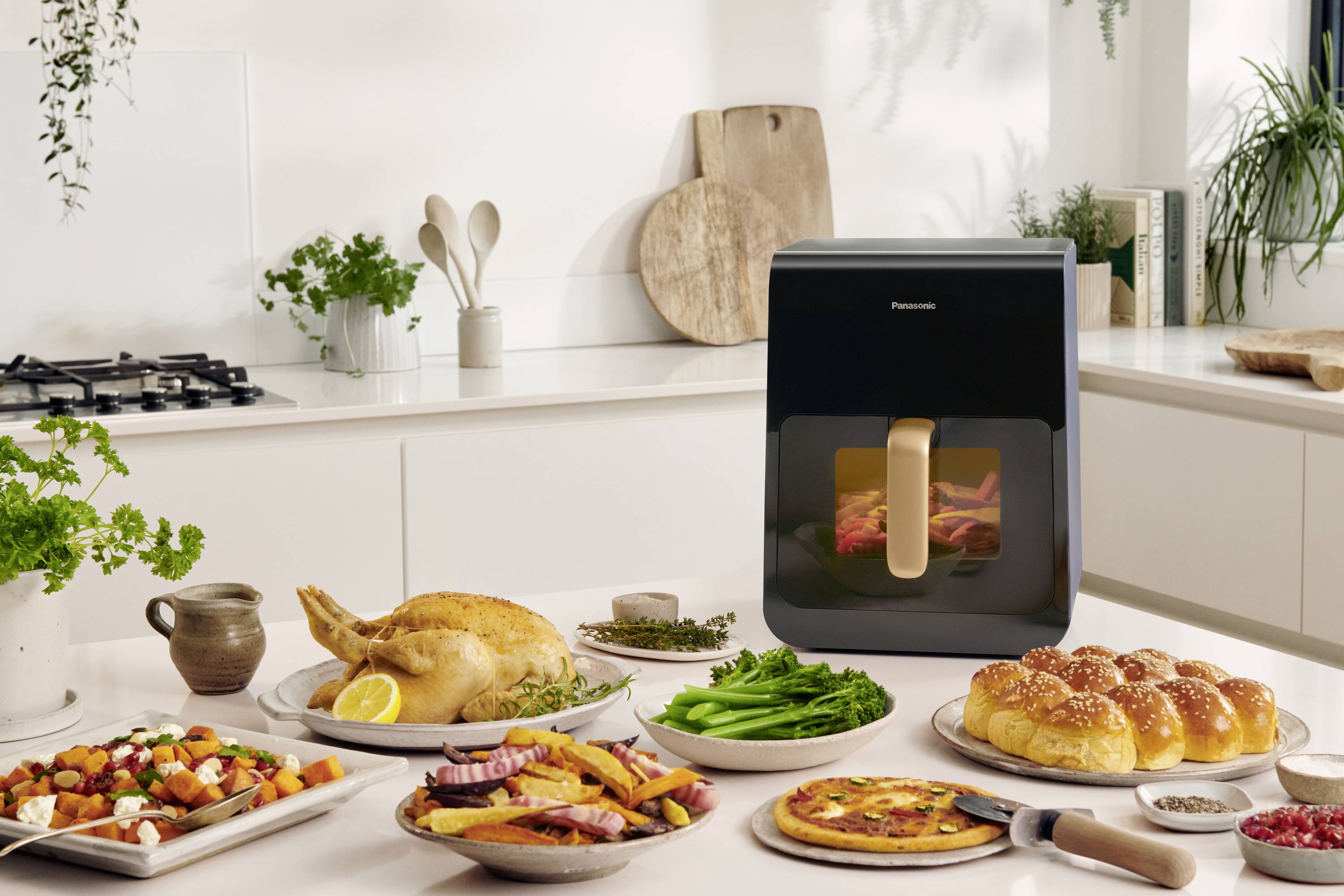 A modern kitchen oven on a worktop, surrounded by various dishes such as roast chicken, vegetables, and bread rolls.