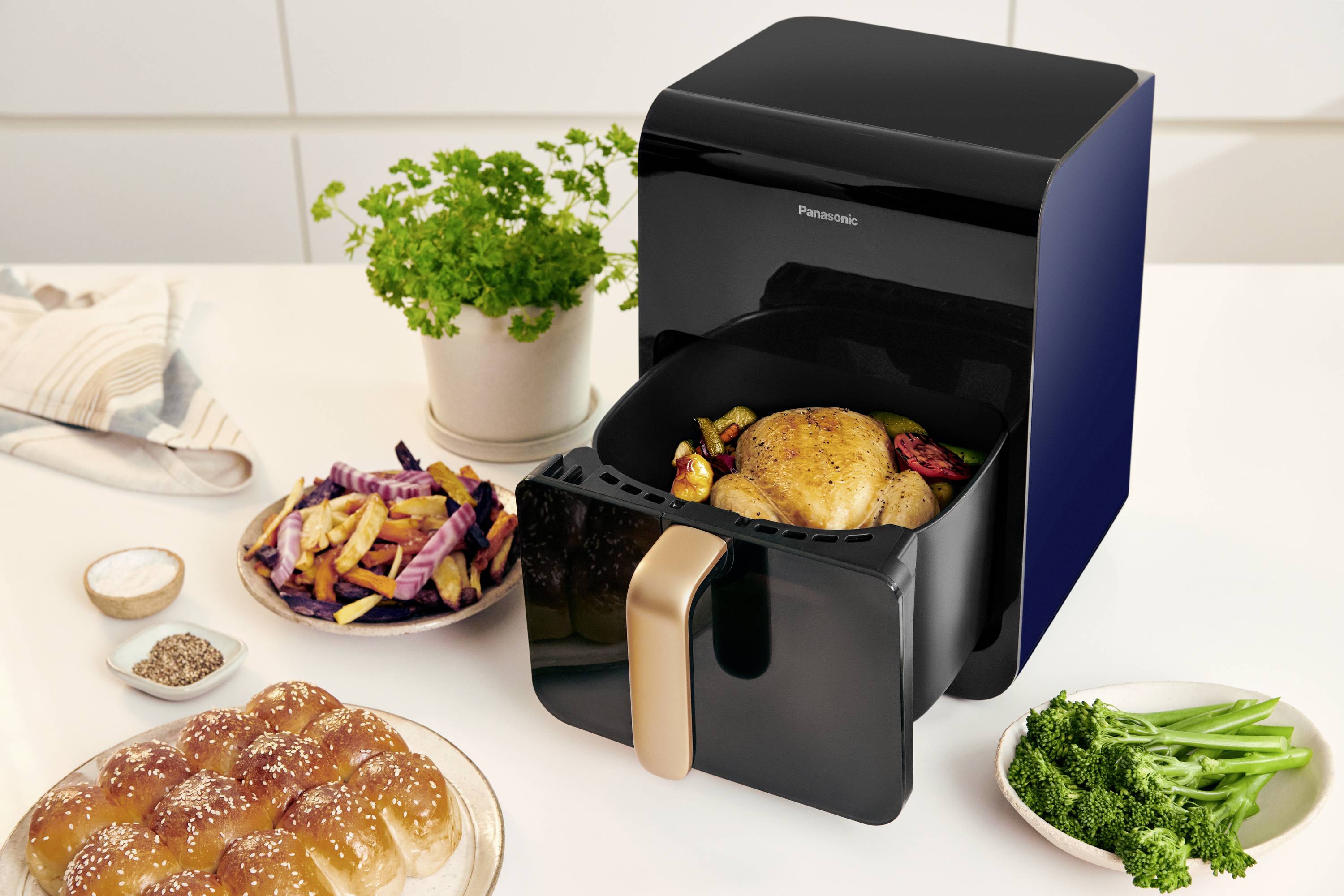 A black air fryer with a golden handle sits on a kitchen worktop. Inside is a roasted chicken with vegetables. Beside it are bread, chips and herbs.