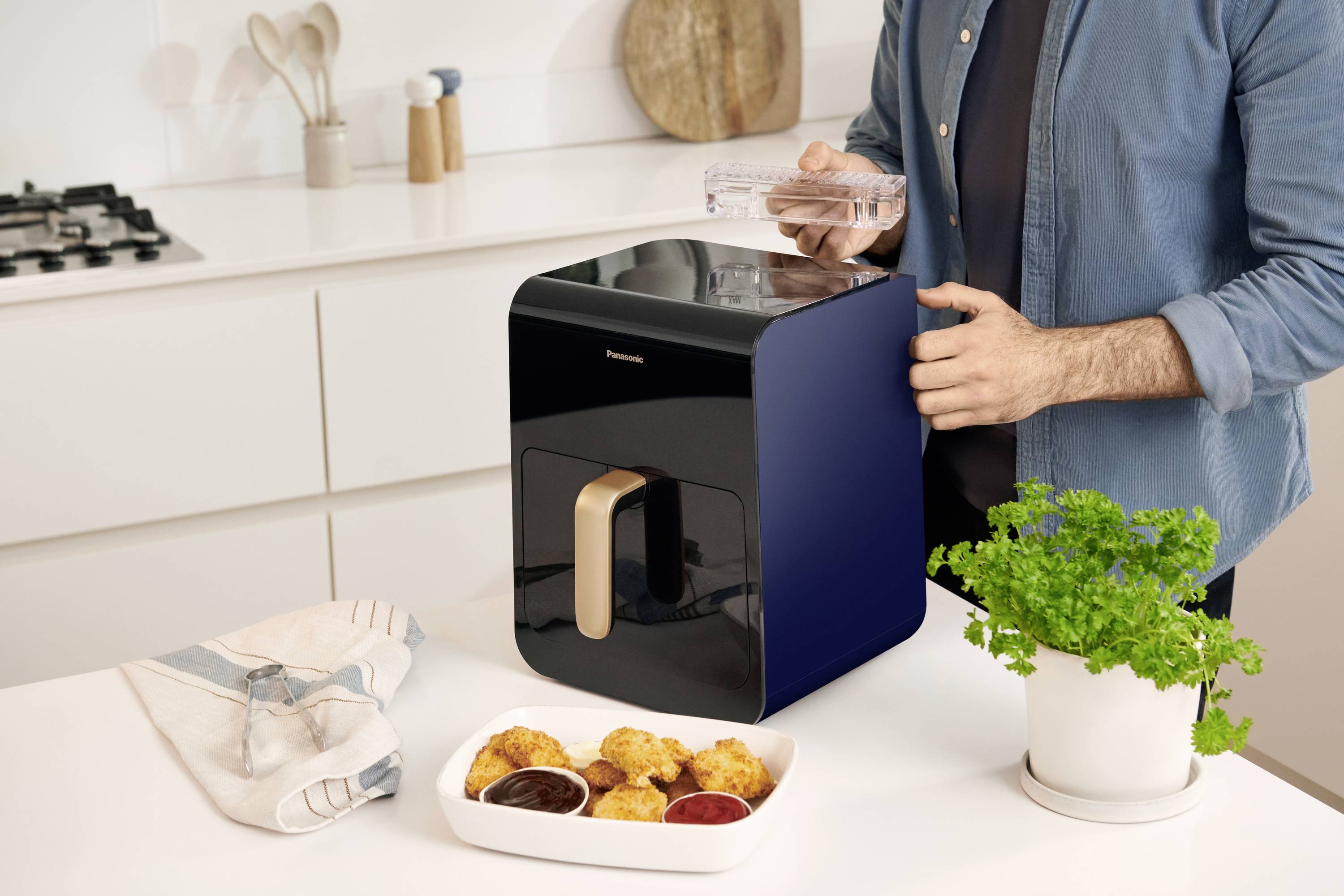 A person is using a modern water filter in a bright kitchen. The table displays serviettes, snacks, and a pot with a green plant.