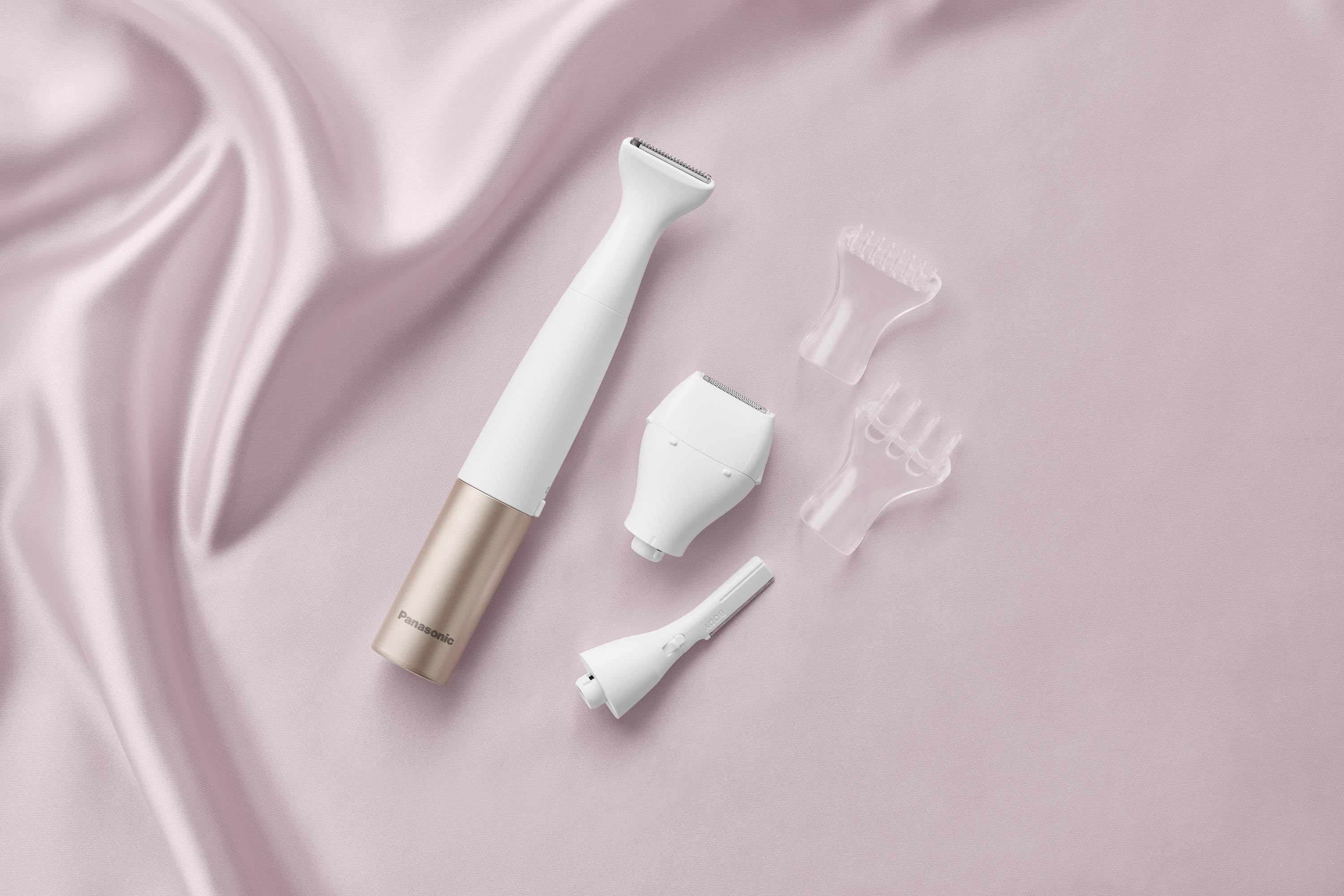 A cordless, electric hair removal device with various attachments lies on a pink silk fabric.