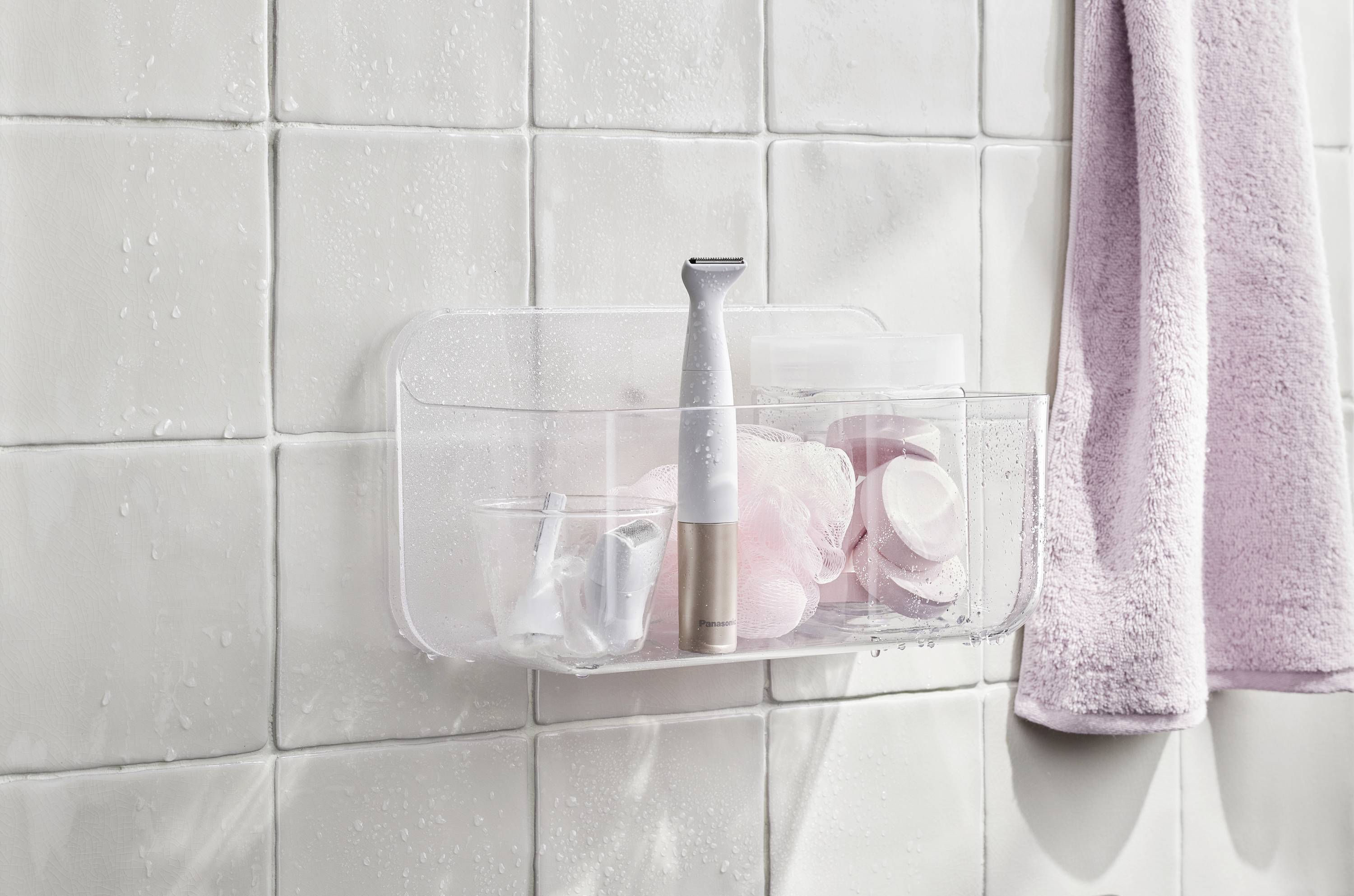 Shower shelf with toiletries and an electric razor, with a pink towel beside it. White tiles in the background.