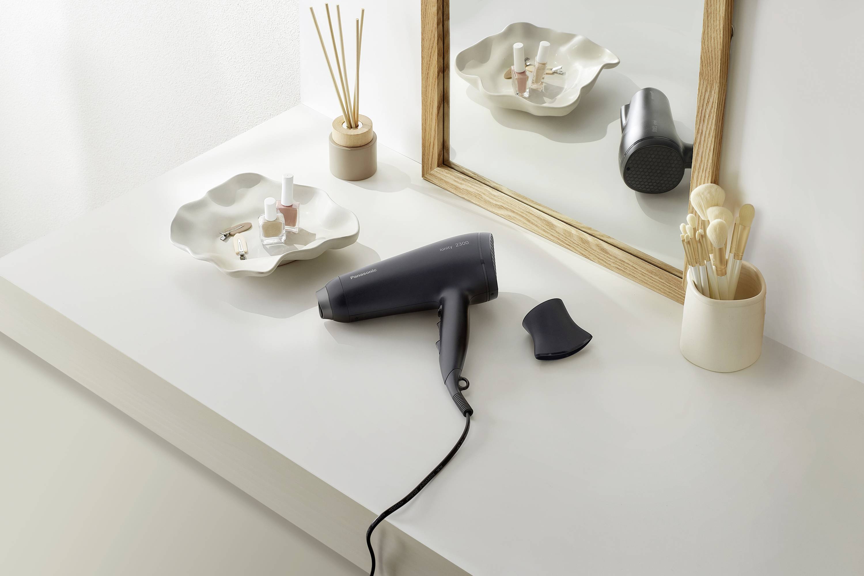 A hairdryer on a white table in front of a mirror. Beside it are brushes, perfume bottles and other accessories. Modern, minimalist aesthetic.