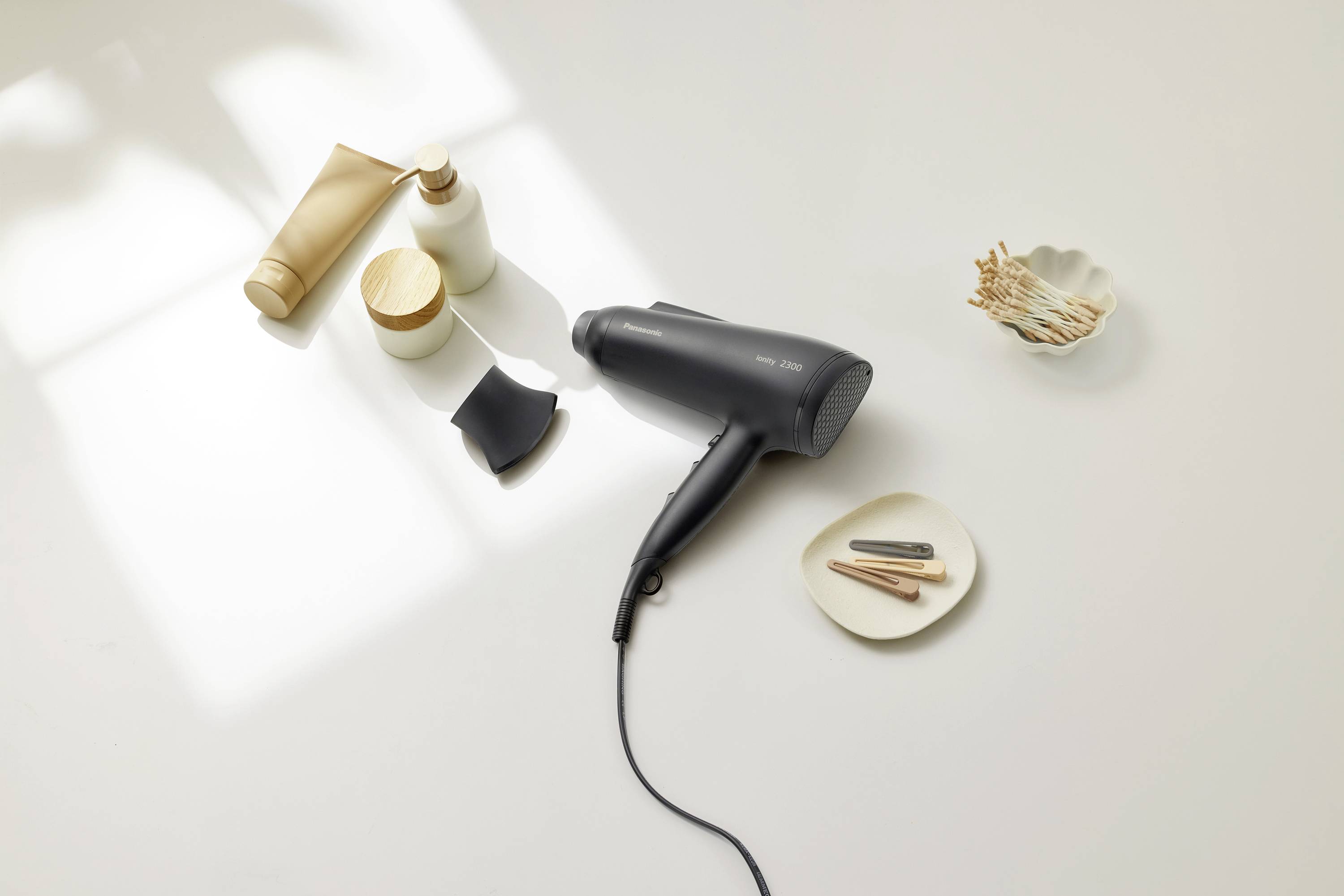 A hairdryer is placed on a table, surrounded by beauty products. Natural light streams through a window, softly illuminating the scene.