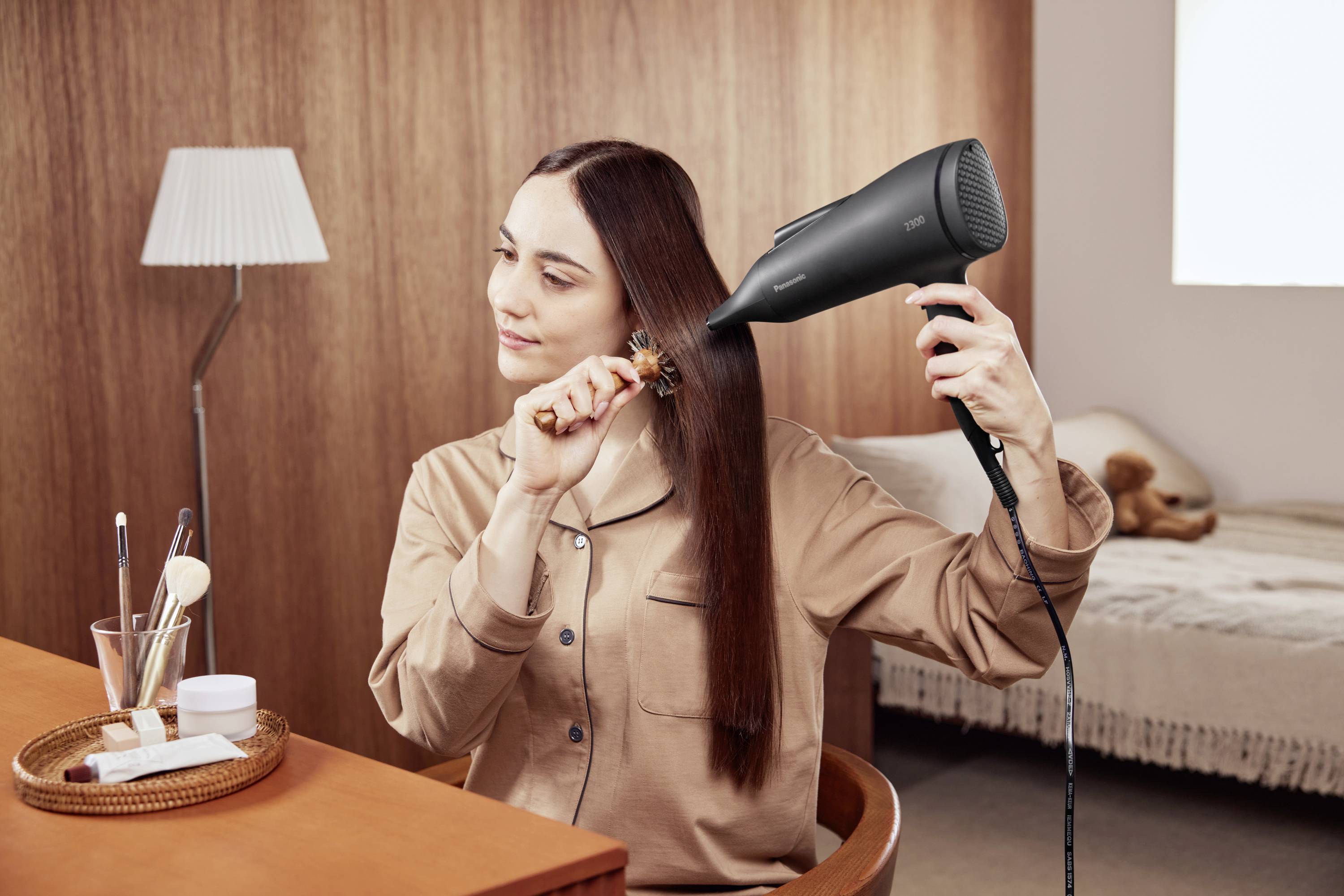 A woman is blow-drying her straight hair with a black hairdryer, seated at a table with make-up accessories in the background.