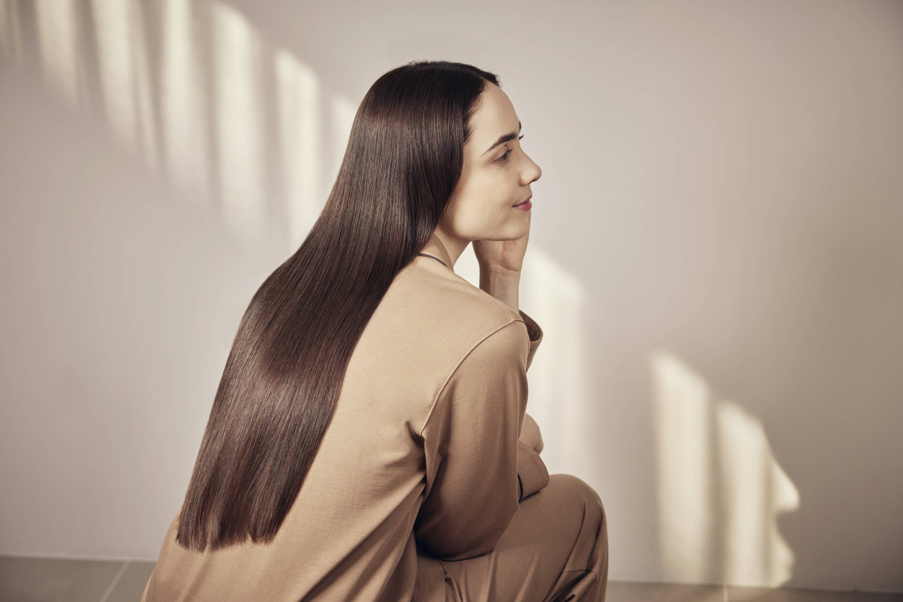 A woman with long, straight brown hair sits in a side-on pose in front of a wall with light and shadow. She is wearing beige clothing.