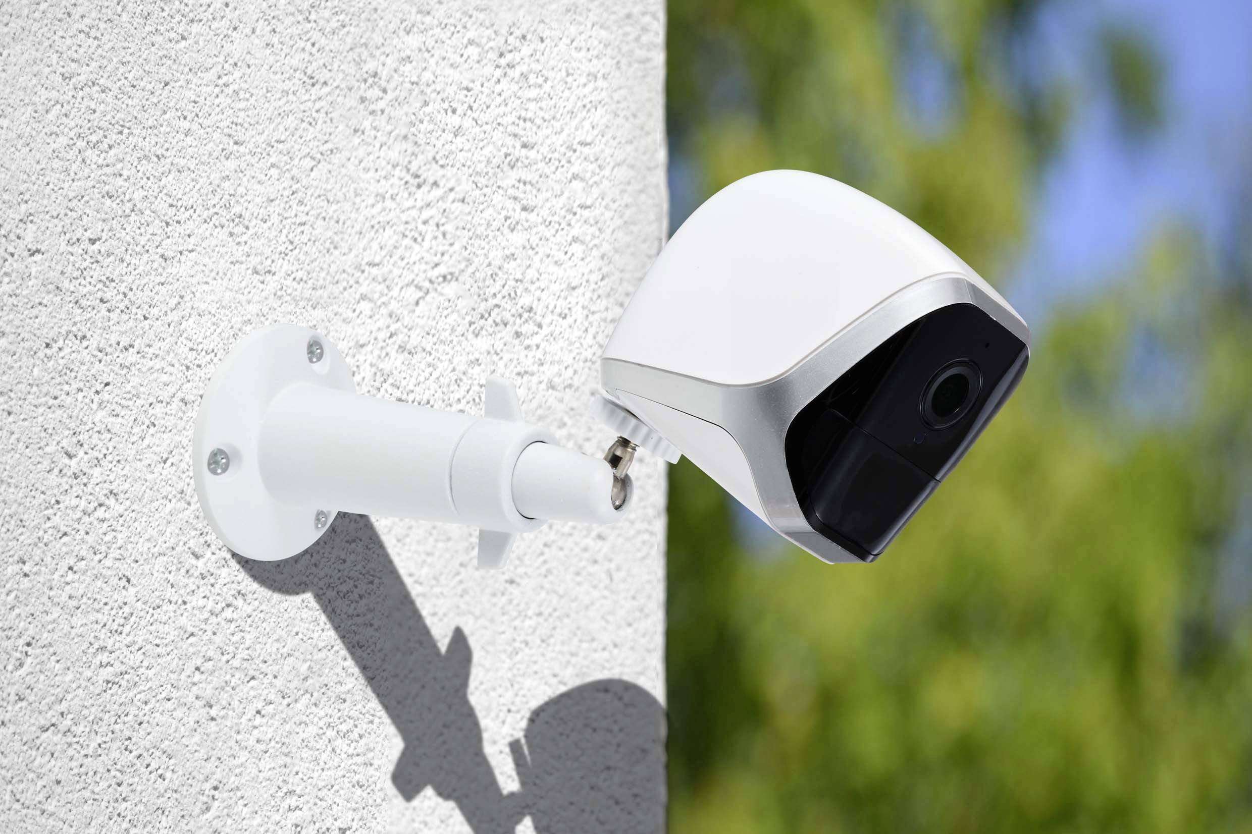 A security camera mounted on a white wall, facing in one direction. The background shows blurred green, possibly plants.
