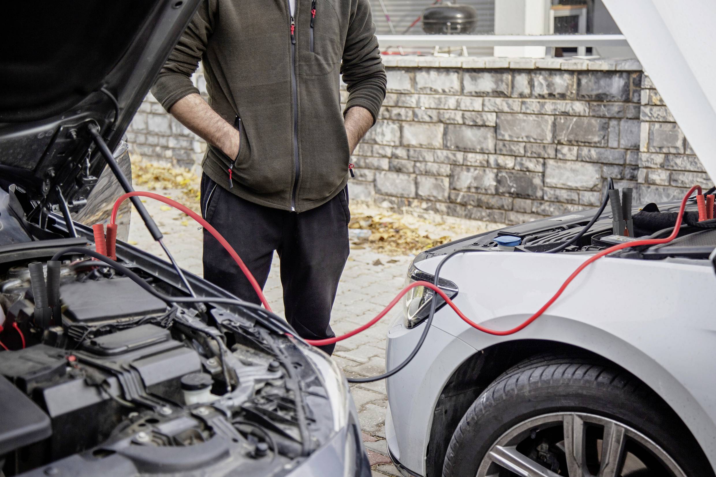A person is jump-starting a car with another vehicle. Both cars have their bonnets open, and red jump leads are connected.