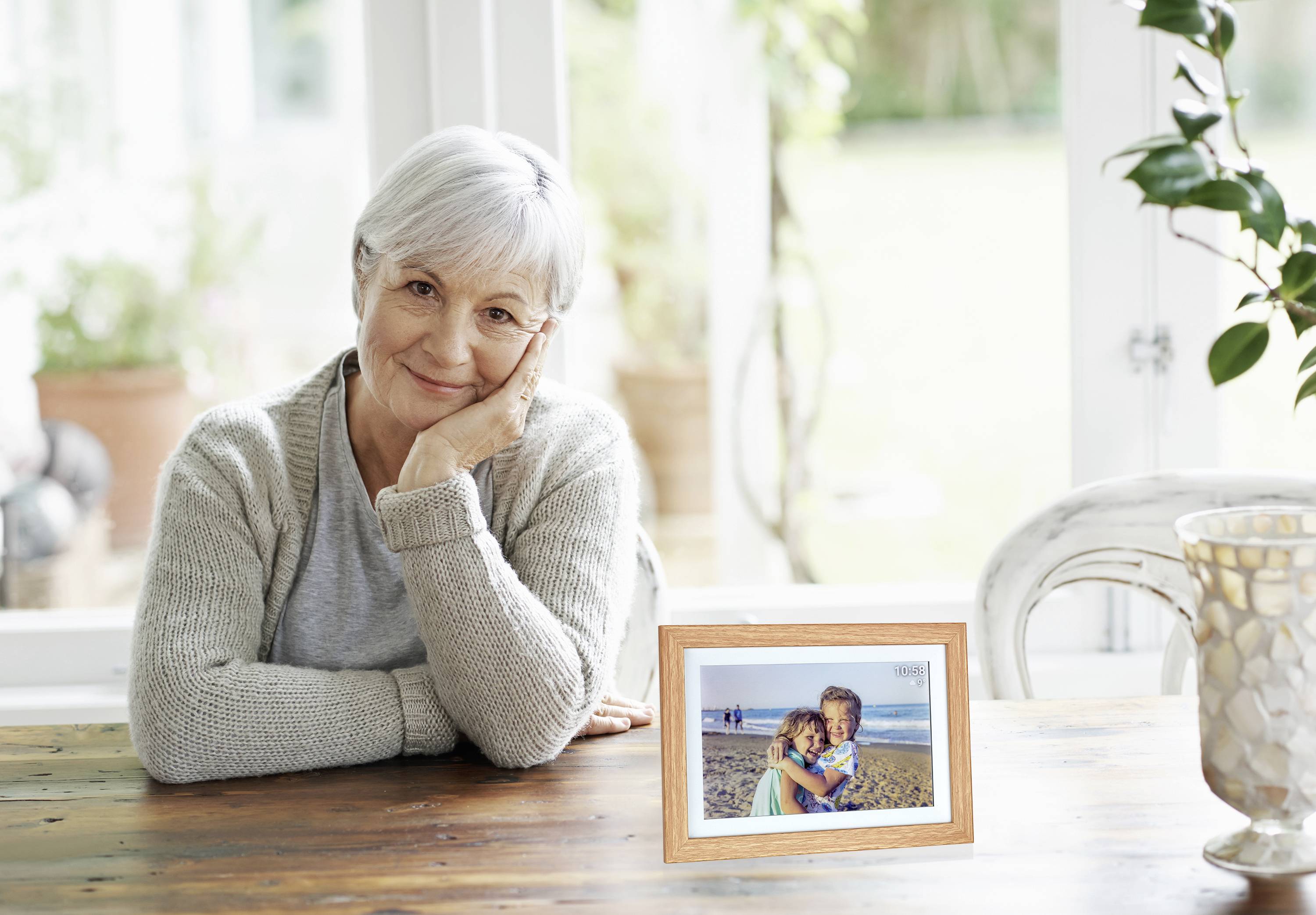 An elderly woman sits smiling at a table in a sunny room, with a framed photograph of two people on the beach beside her.