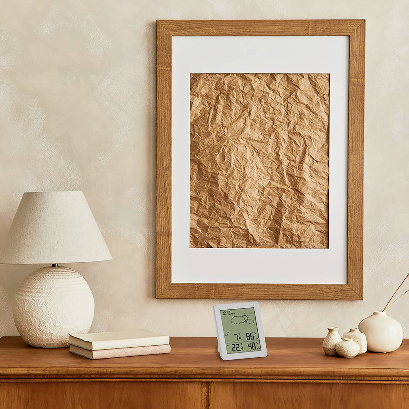 A wooden table with a lamp, two books, a digital thermometer, and small ceramics. On the wall hangs a picture with brown paper.