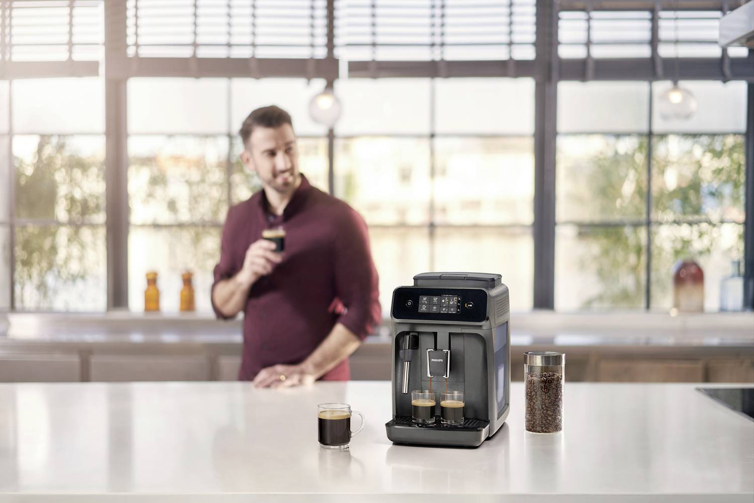 A man is holding a cup of coffee and standing behind a modern coffee machine on a kitchen worktop.