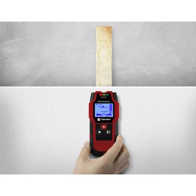 A hand holds an electronic measuring device against a wall. The display shows a diagram for detecting a wooden beam behind the wall.