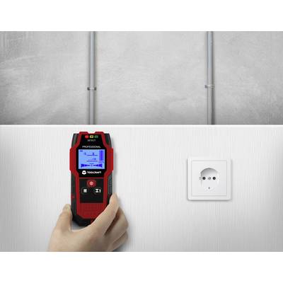 A person is holding a digital cable detector in front of a white wall. A plug socket is visible to the right of the device.