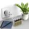 A plug socket with a USB charging adapter is charging four AA batteries. Next to it, a green plant in a pot and a notebook sit on the table.