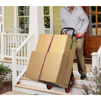 A man is transporting two large boxes up the steps of a veranda on a trolley. A house is visible in the background.
