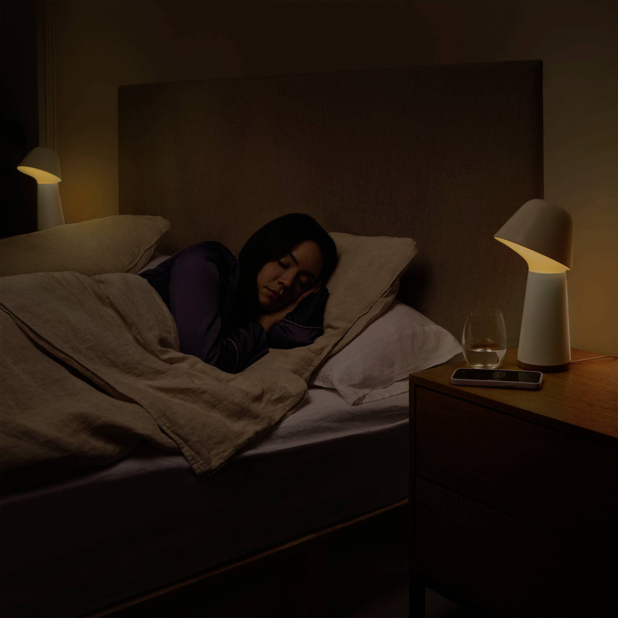 A woman is sleeping in a darkened bedroom, snuggled under a blanket. A glass of water sits next to a lamp on the bedside table.