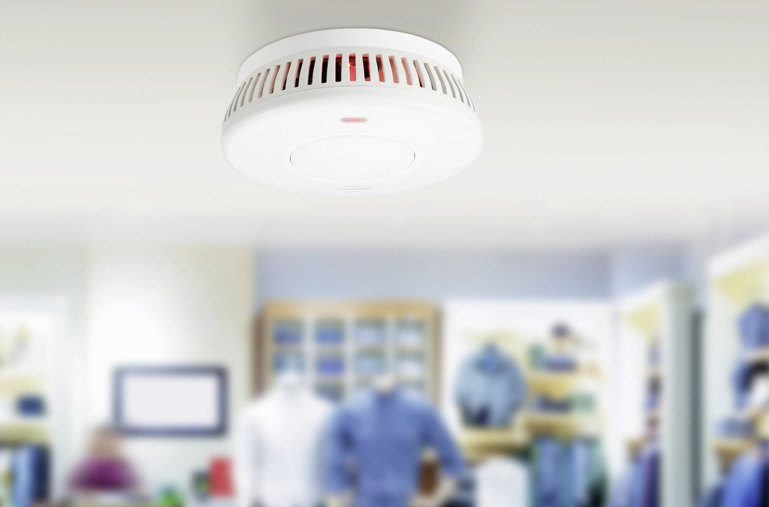 A smoke detector is mounted on the ceiling of a modern commercial space. In the background, clothing shelves are blurred out of focus.