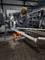 A worker in protective clothing is drilling into a metal part held in an orange clamp within a workshop environment.