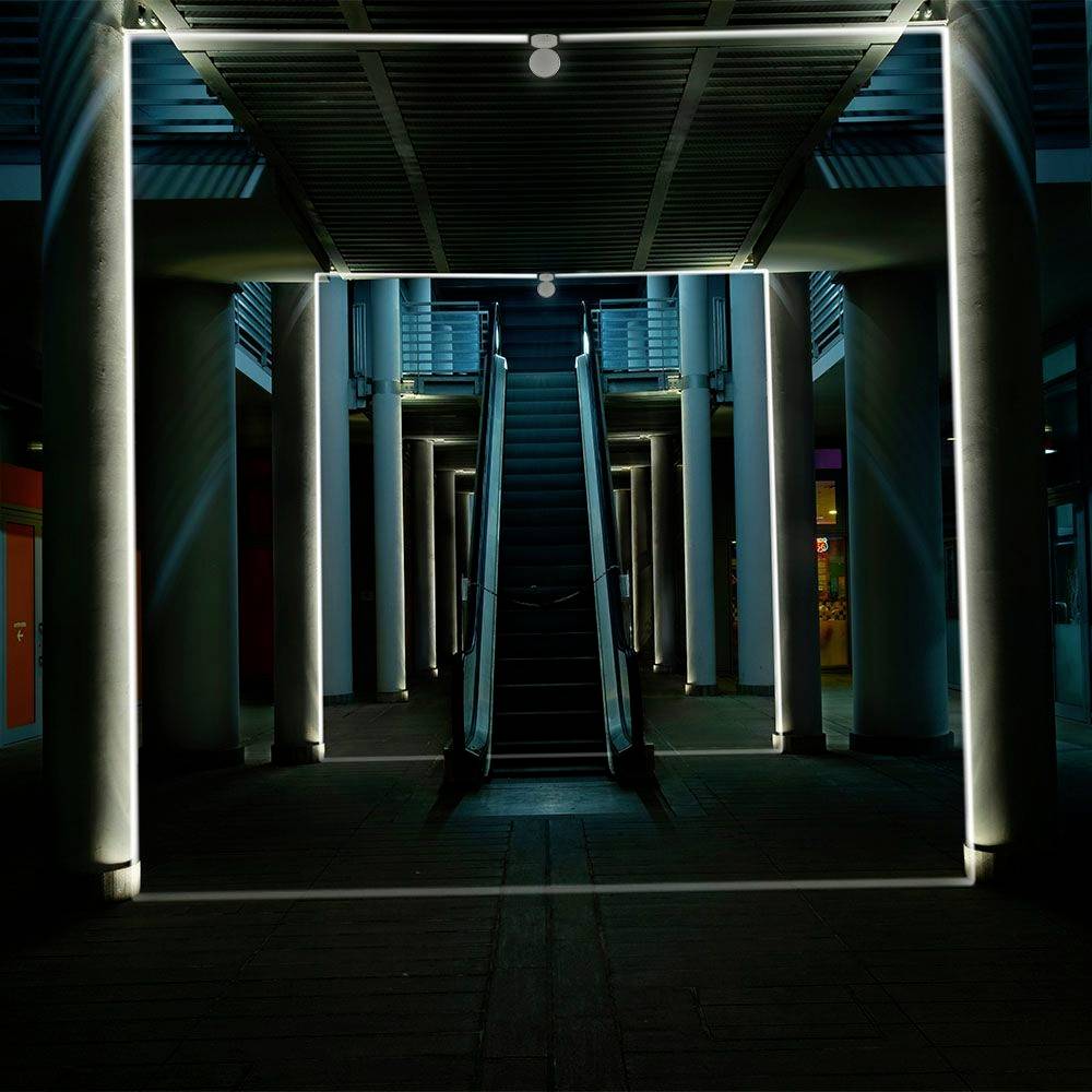 A modern, illuminated entrance with an escalator in an urban area, surrounded by columns, photographed at night.