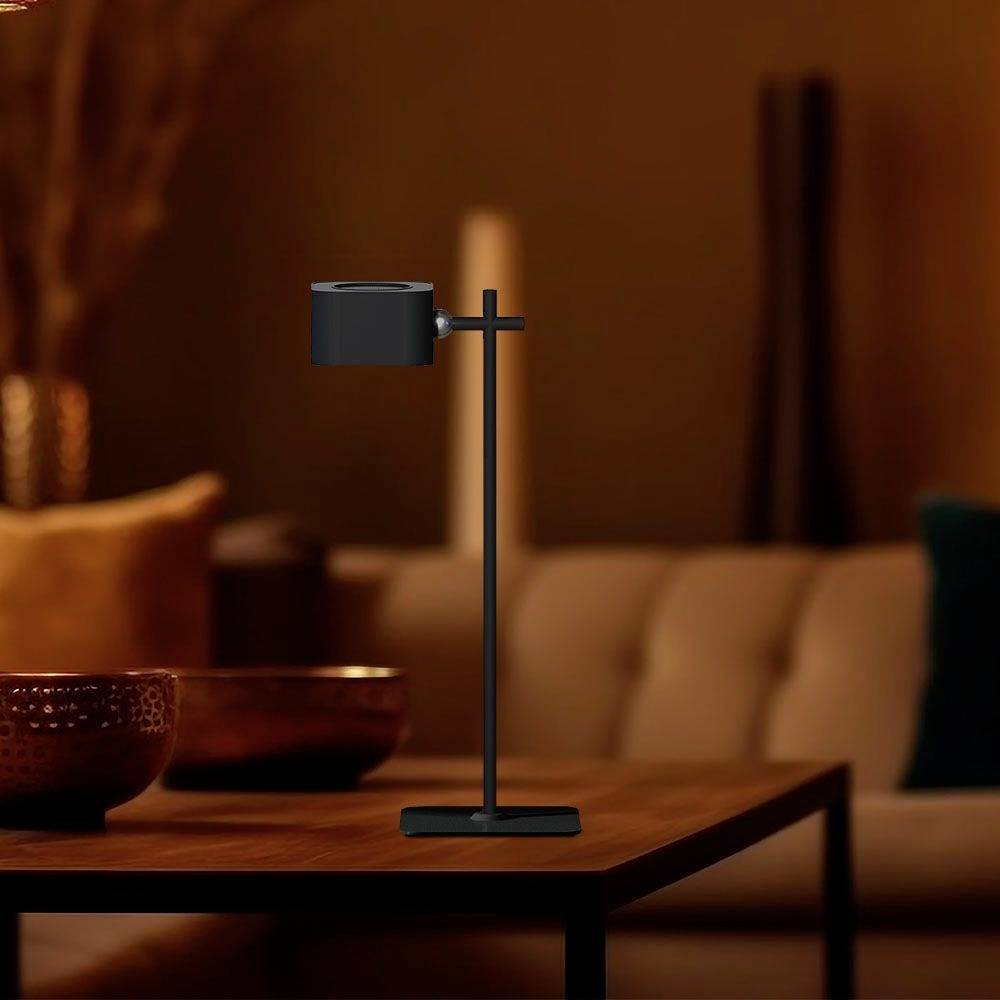 A modern table lamp with an adjustable lampshade sits on a wooden table in a cosily lit room with a blurred sofa in the background.