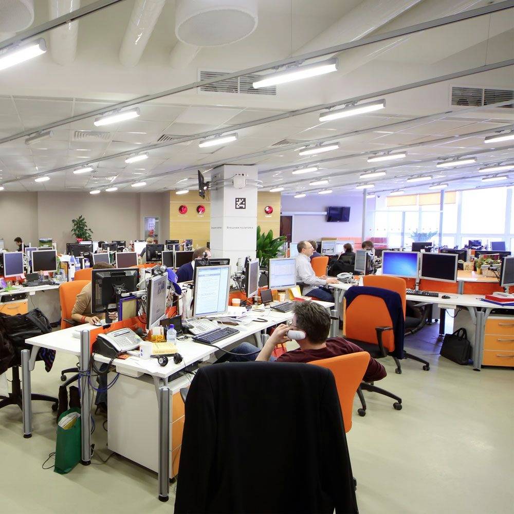 An open-plan office with multiple desks, computers, and people working. Bright lighting and large windows in the background.