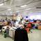 An open-plan office with multiple desks, computers, and people working. Bright lighting and large windows in the background.