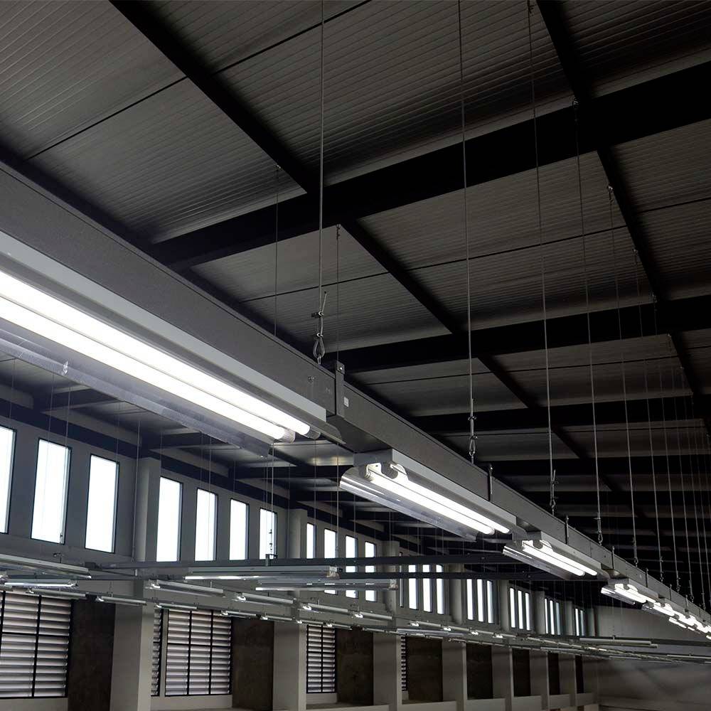 Illuminated ceiling lights in a large warehouse or factory hall with visible metal beams and windows along the walls.