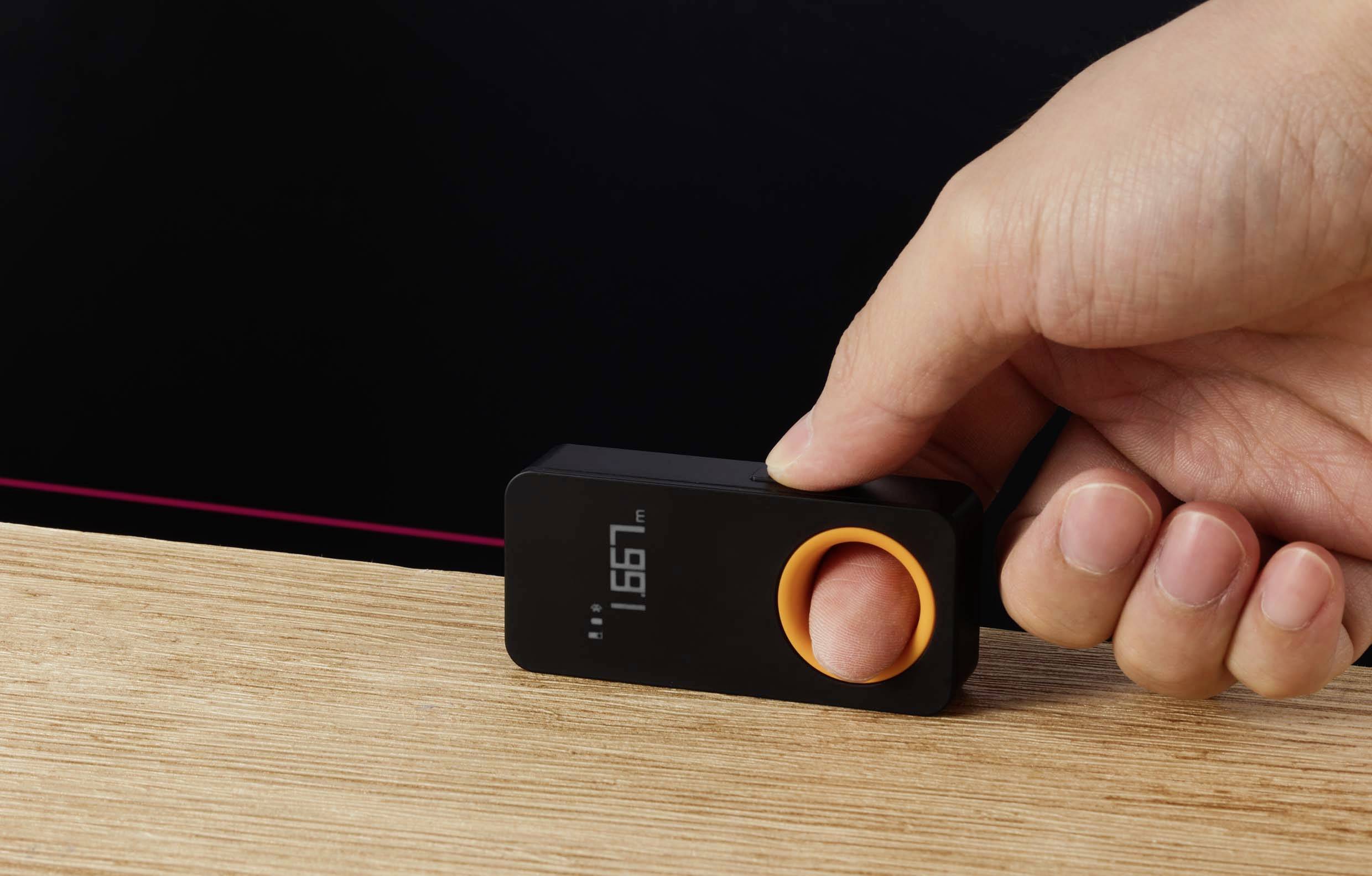 A hand presses a digital measuring device with a display onto a wooden table, with the result shown on the screen.