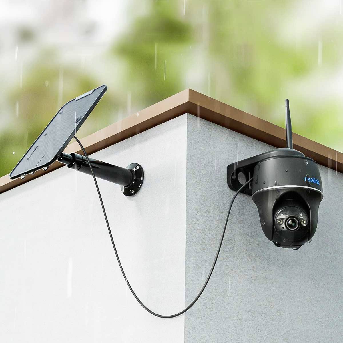 A security camera with a solar panel is mounted on the corner of a building. It is raining and the camera is pointed downwards.