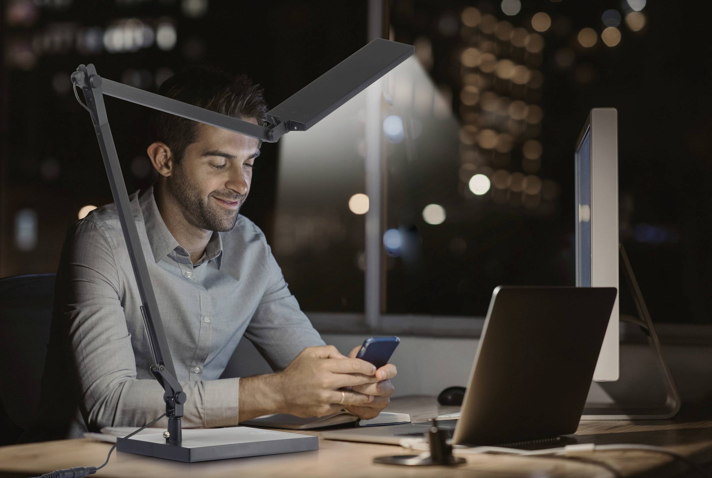 A man sits in an office in the evening at a desk, working on a computer and glancing at his mobile phone, while being illuminated by a lamp.