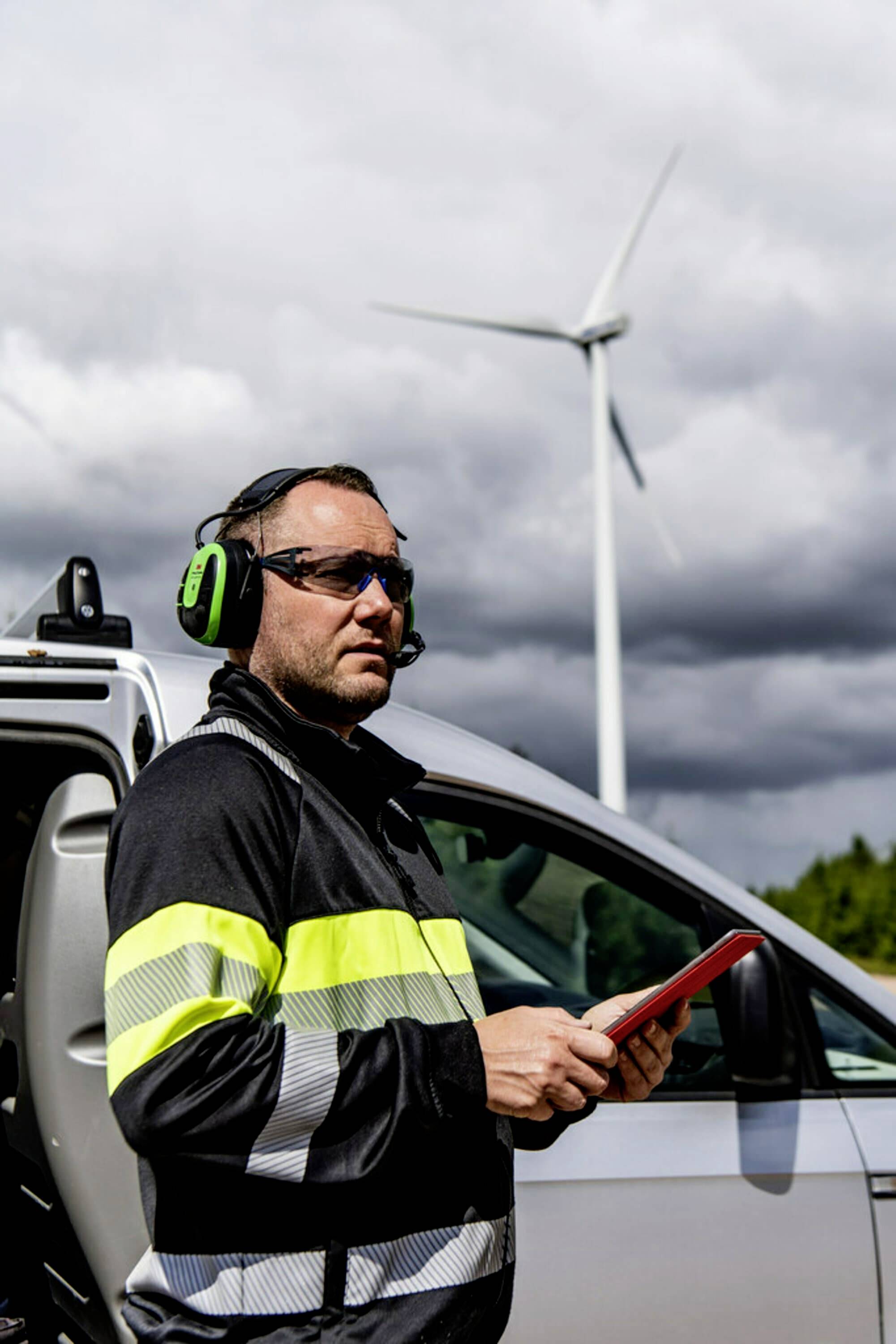 A technician wearing protective clothing and headphones is standing next to a delivery van and holding a tablet; a wind turbine is visible in the background.