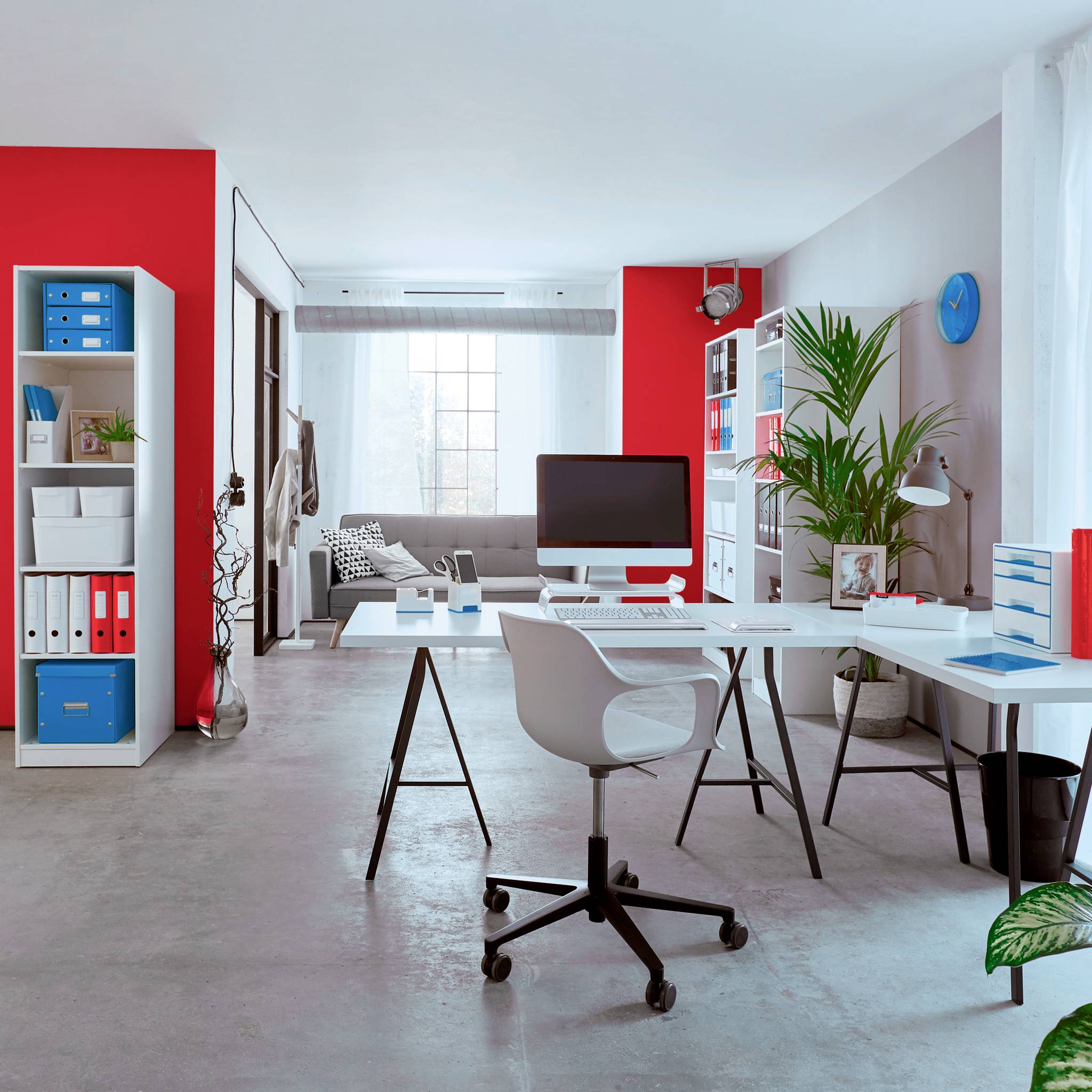 Bright office with a white desk, red bookshelf, computer, office chair and grey sofa in the background. Plants decorate the room.