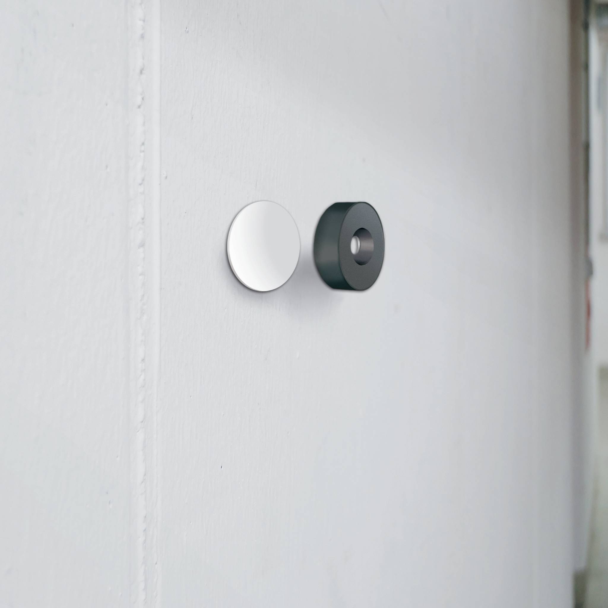 Two differently shaped wall hooks are mounted on a white wall. The left hook is round and silver-coloured, the right one is cylindrical and black.