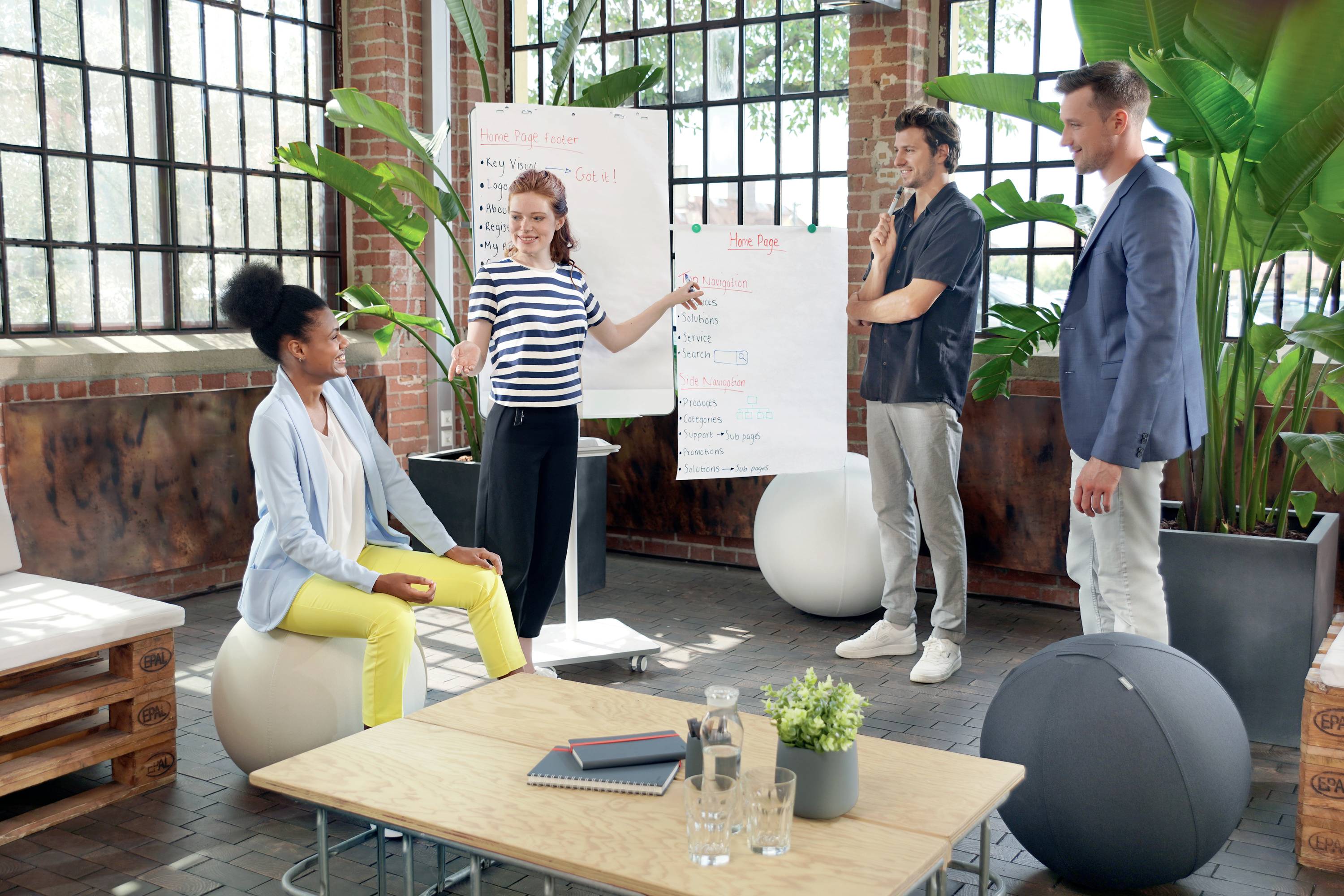 People in a modern office discussing a project; a woman is presenting notes on a flipchart while the others listen.