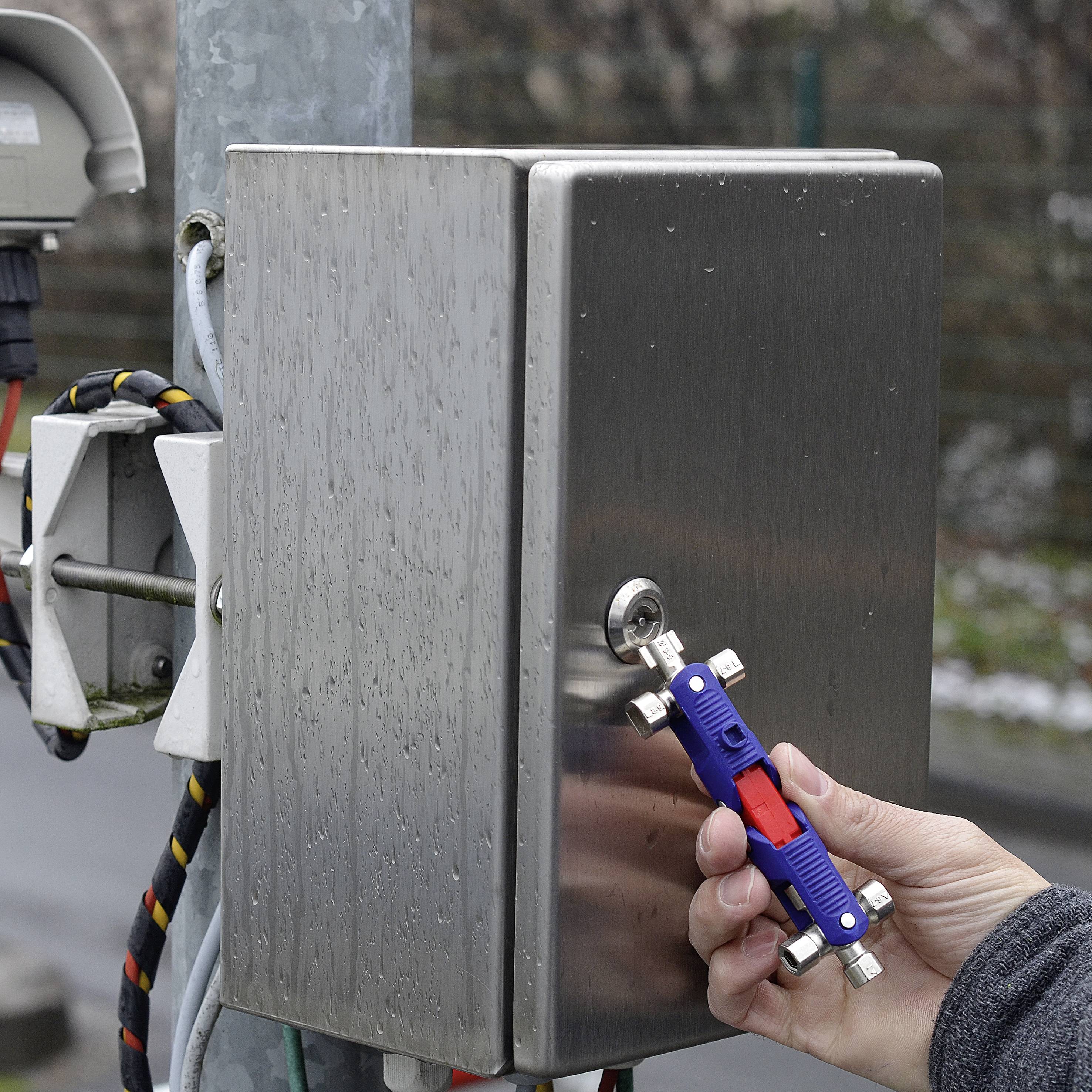 'Hand holding multiple keys, opens silver metal switch box on a pillar with cables. Outside, overcast weather.'