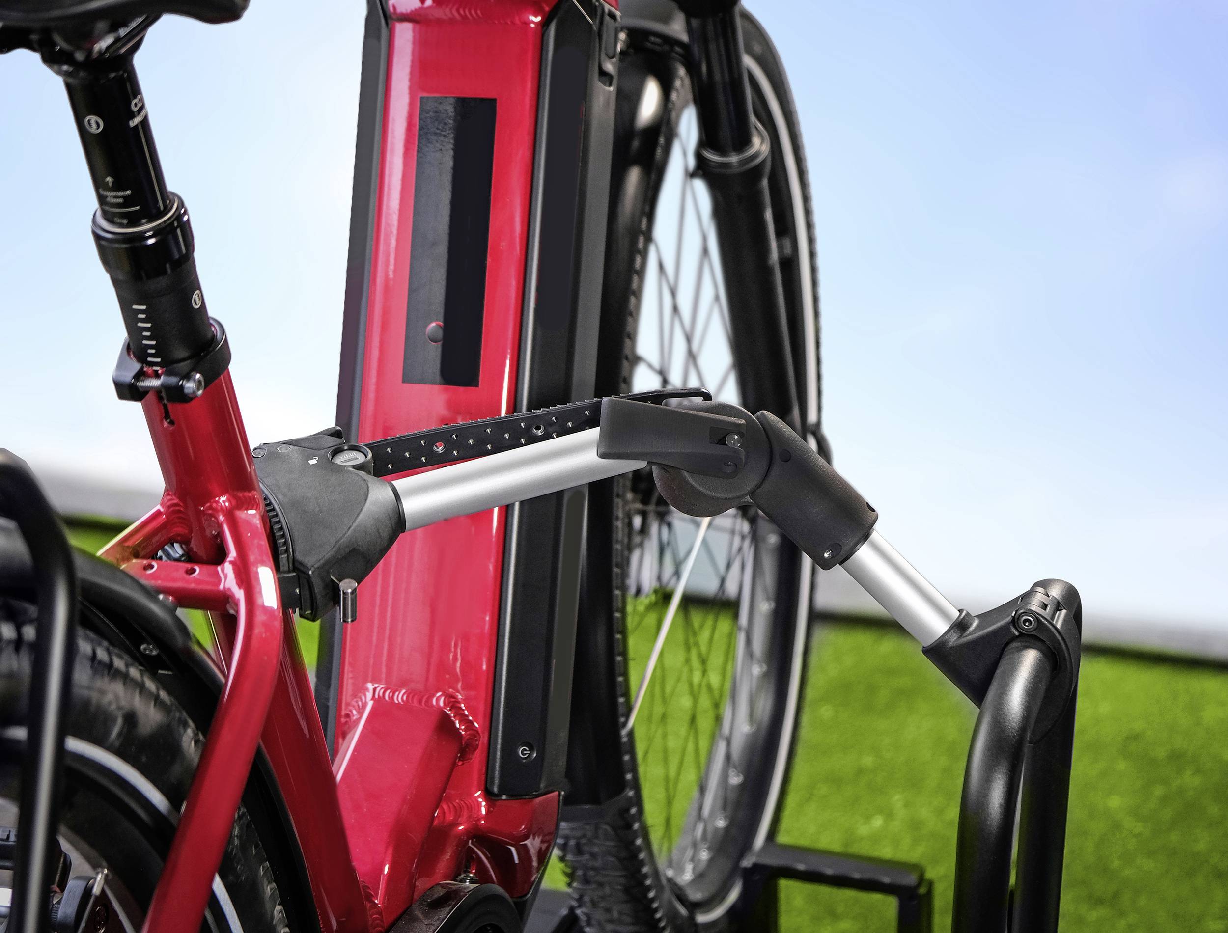 A red e-bike is mounted on a stand against a green background with a blue sky. Detailed view of the battery and frame.