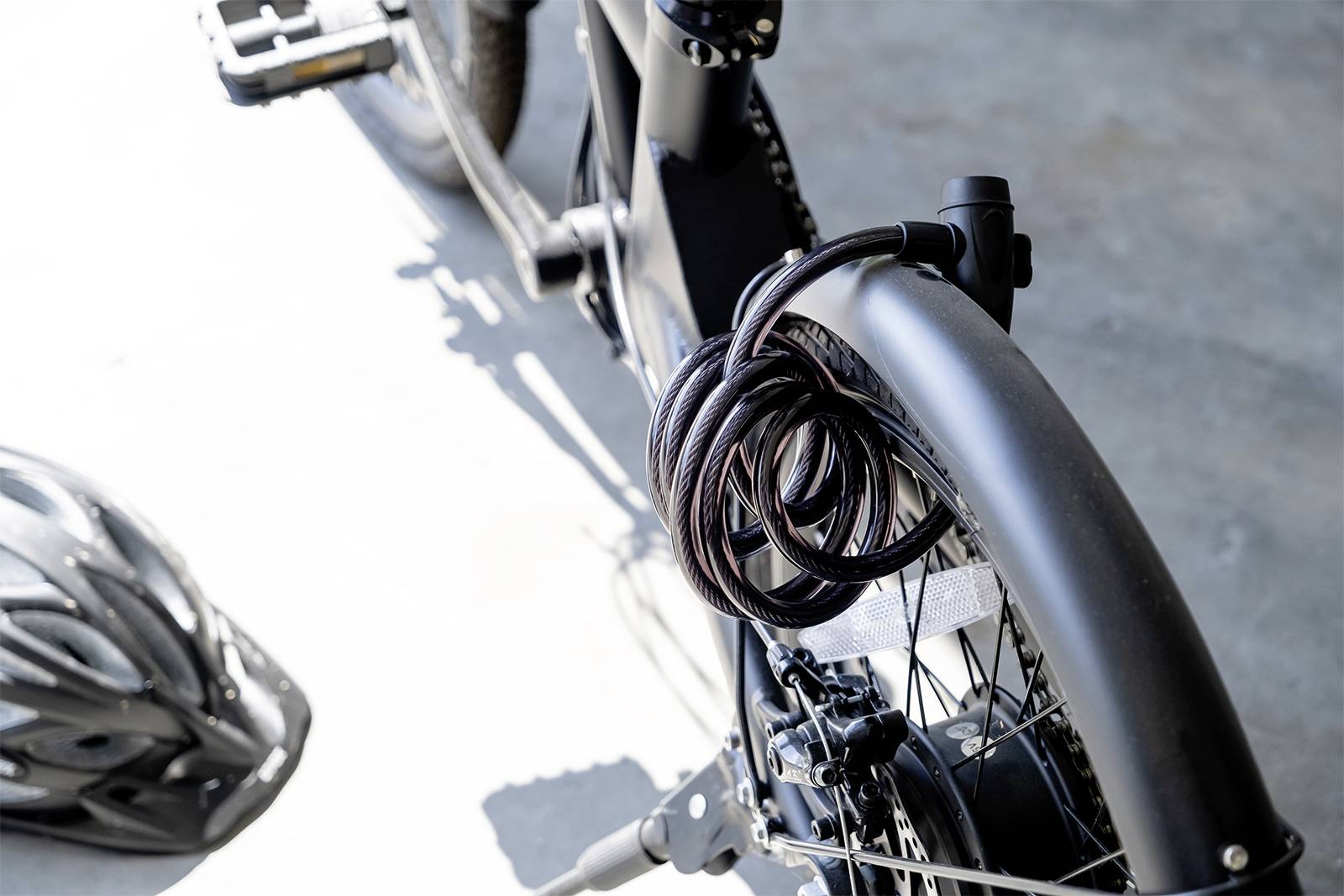 Bicycle with a cable lock attached, mounted on a bike rack, with a bicycle helmet beside it, photographed in daylight.