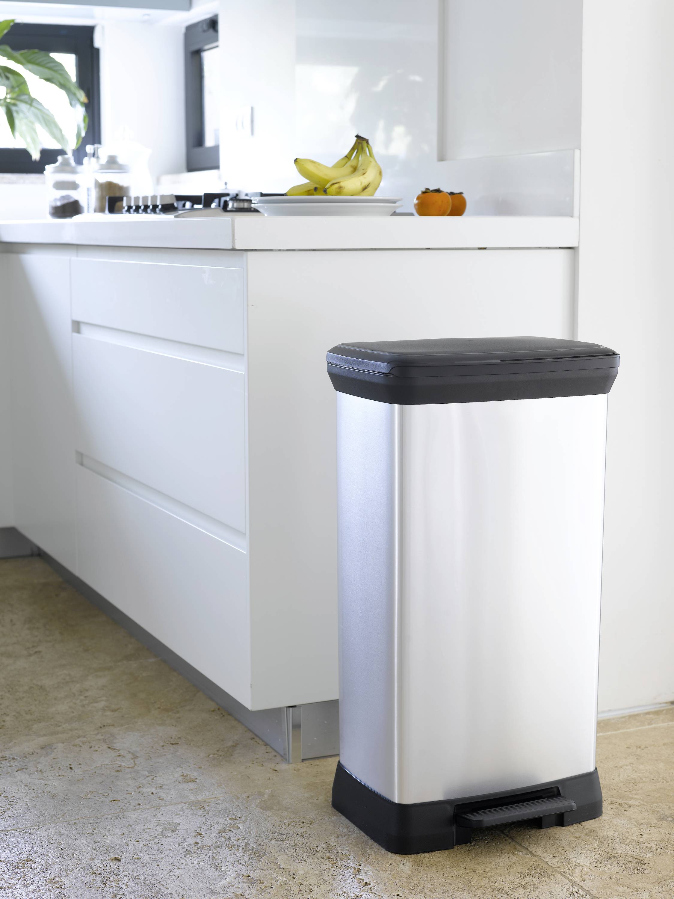 Silver pedal bin stands in a light, modern kitchen. Bananas and an orange are lying on the worktop.