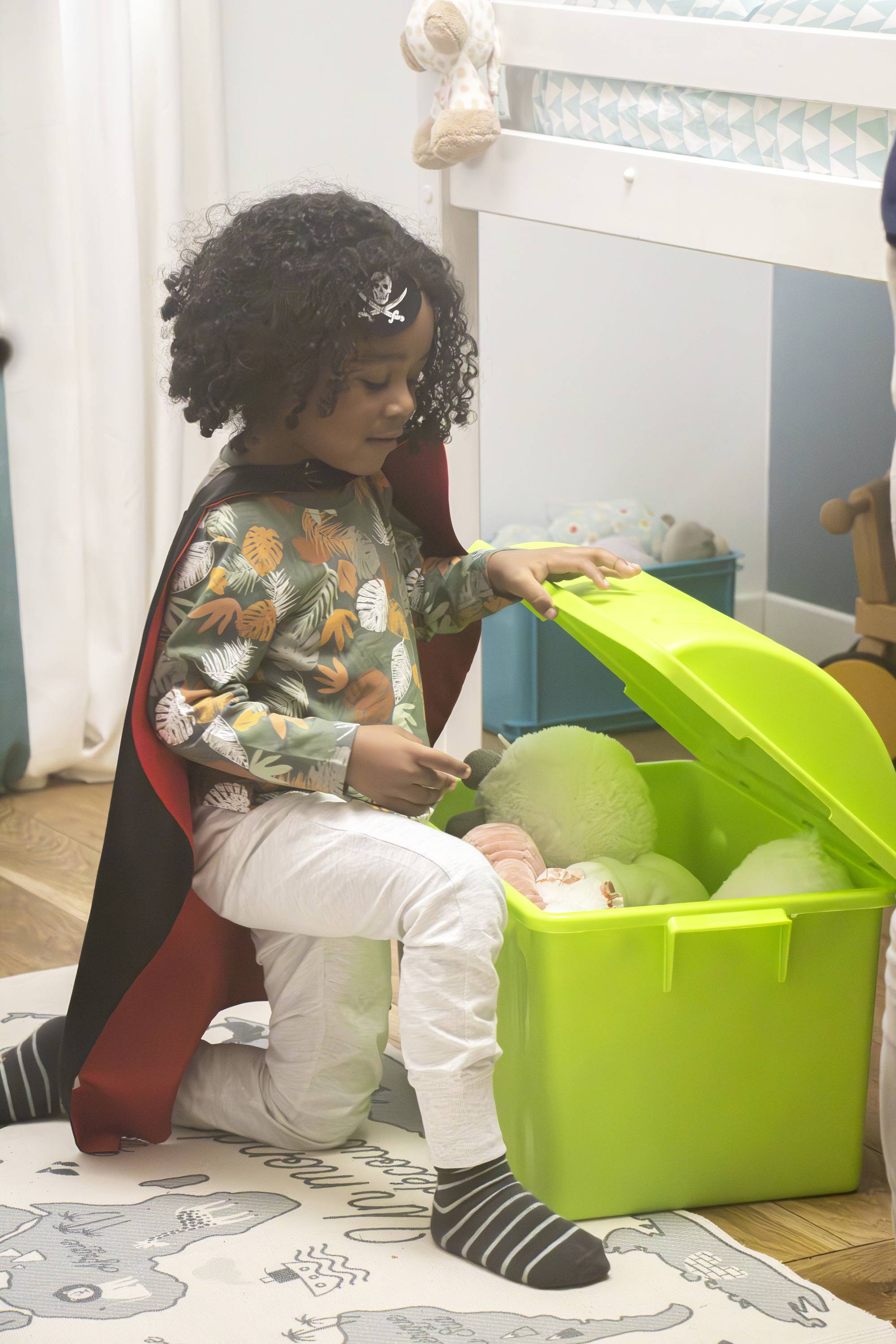 A child wearing a pirate hat plays in a children's bedroom, kneeling in front of an open green toy chest and looking inside.