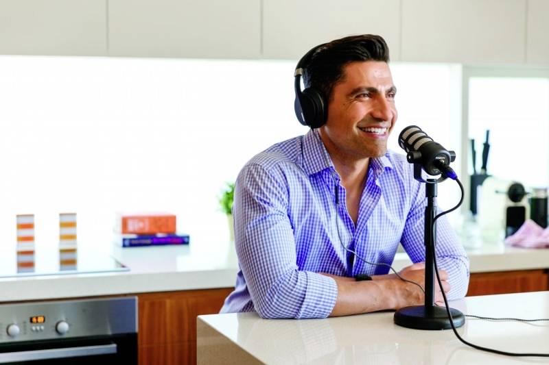 A man sits in a modern kitchen, wearing headphones and speaking into a microphone, smiling. He could be recording a podcast.