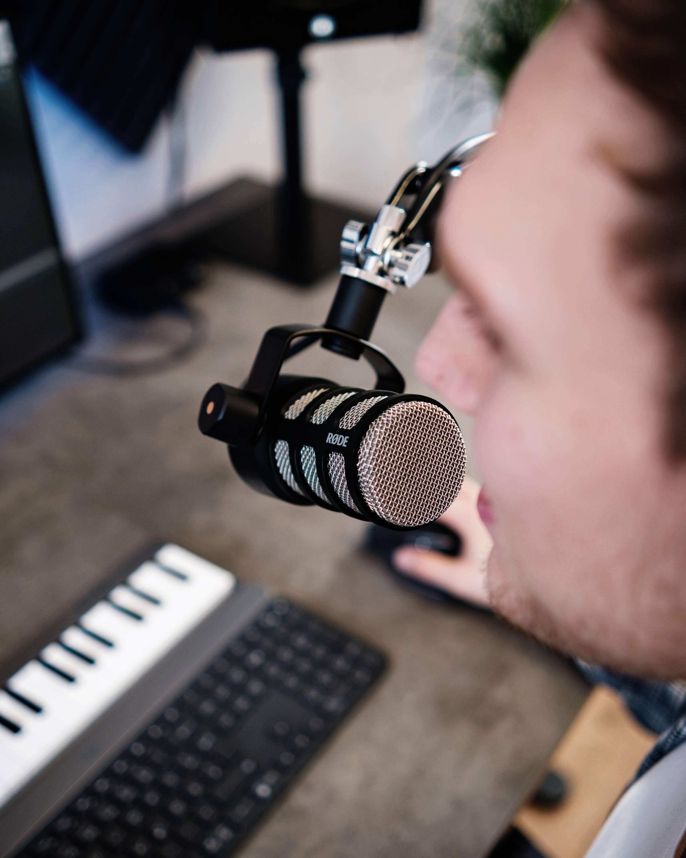 A person is speaking into a microphone in a studio. In the foreground, a computer keyboard and a MIDI keyboard are visible.