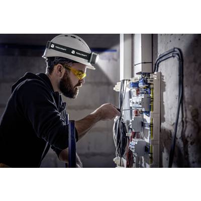 An electrician wearing a safety helmet and protective goggles is working on an open electrical cabinet, checking and adjusting the electrical connections.