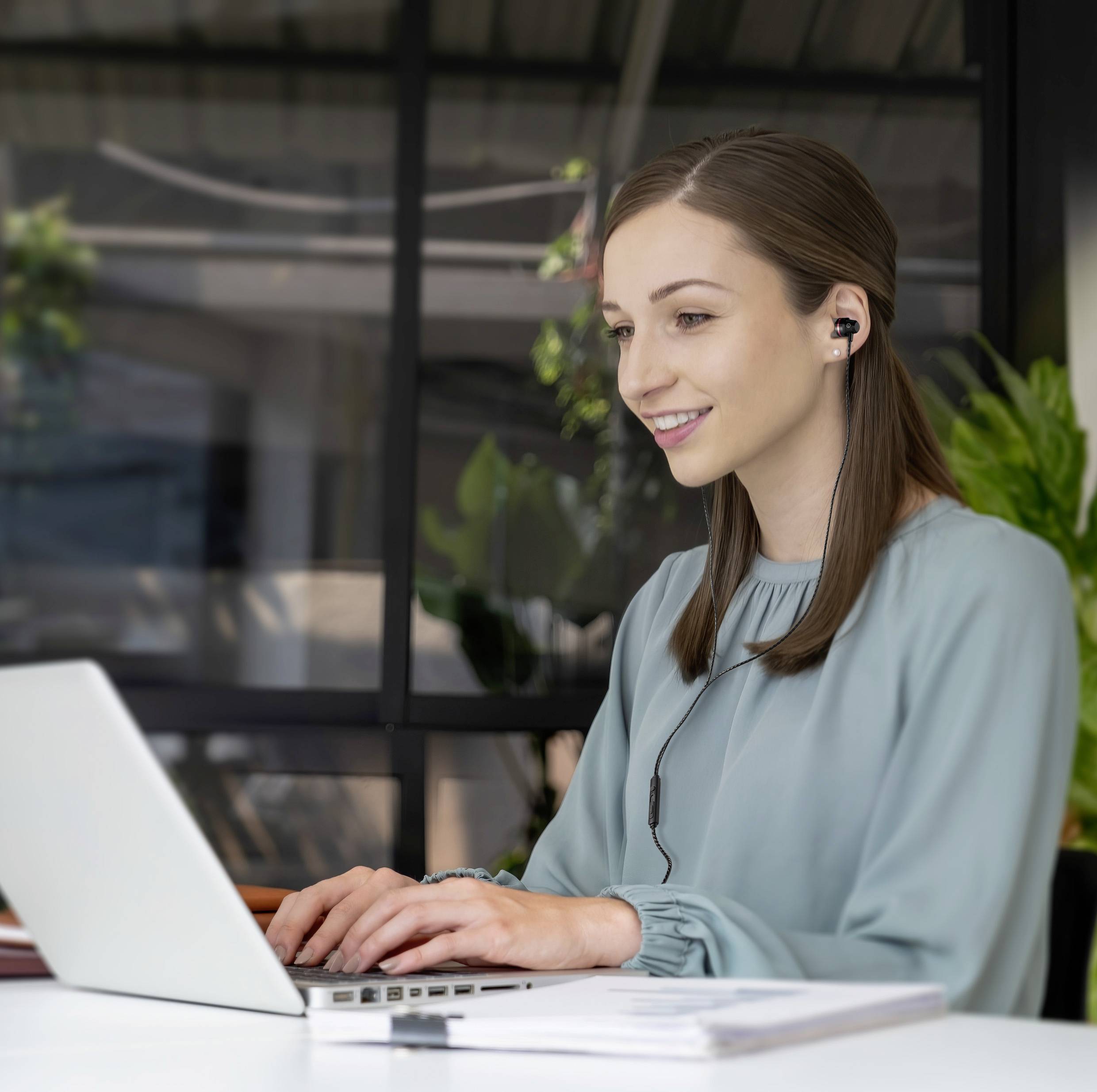 Woman in the office smiles and wears headphones while looking at a laptop. Plants and windows are visible in the background.