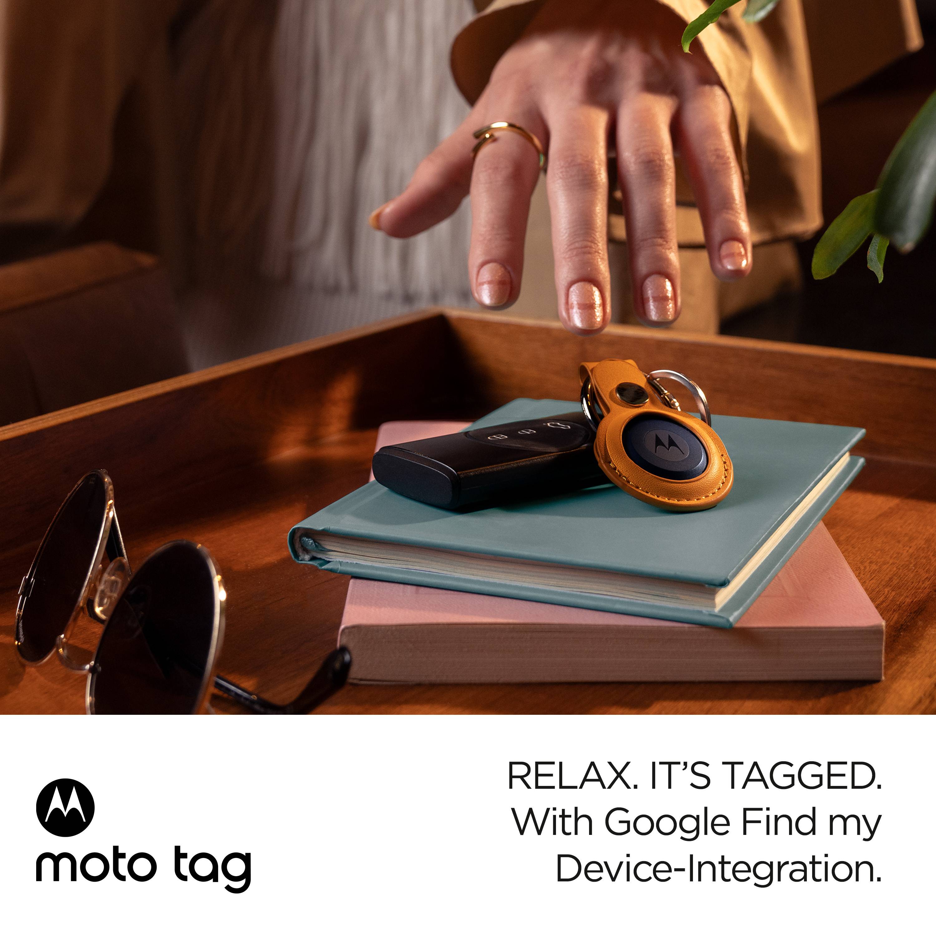 A person places a "moto tag" tracker on a book next to a pair of glasses. The text describes integration with Google Find My Device.<br><br>(Note: The translation is already in British English, so no significant changes were needed.)
