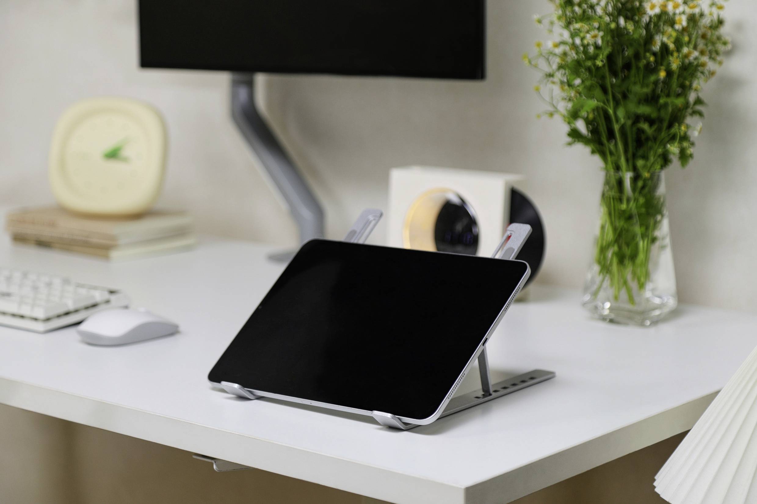 A tablet on a stand is placed on a white desk next to a keyboard and mouse, with a monitor, clock, and vase of flowers in the background.